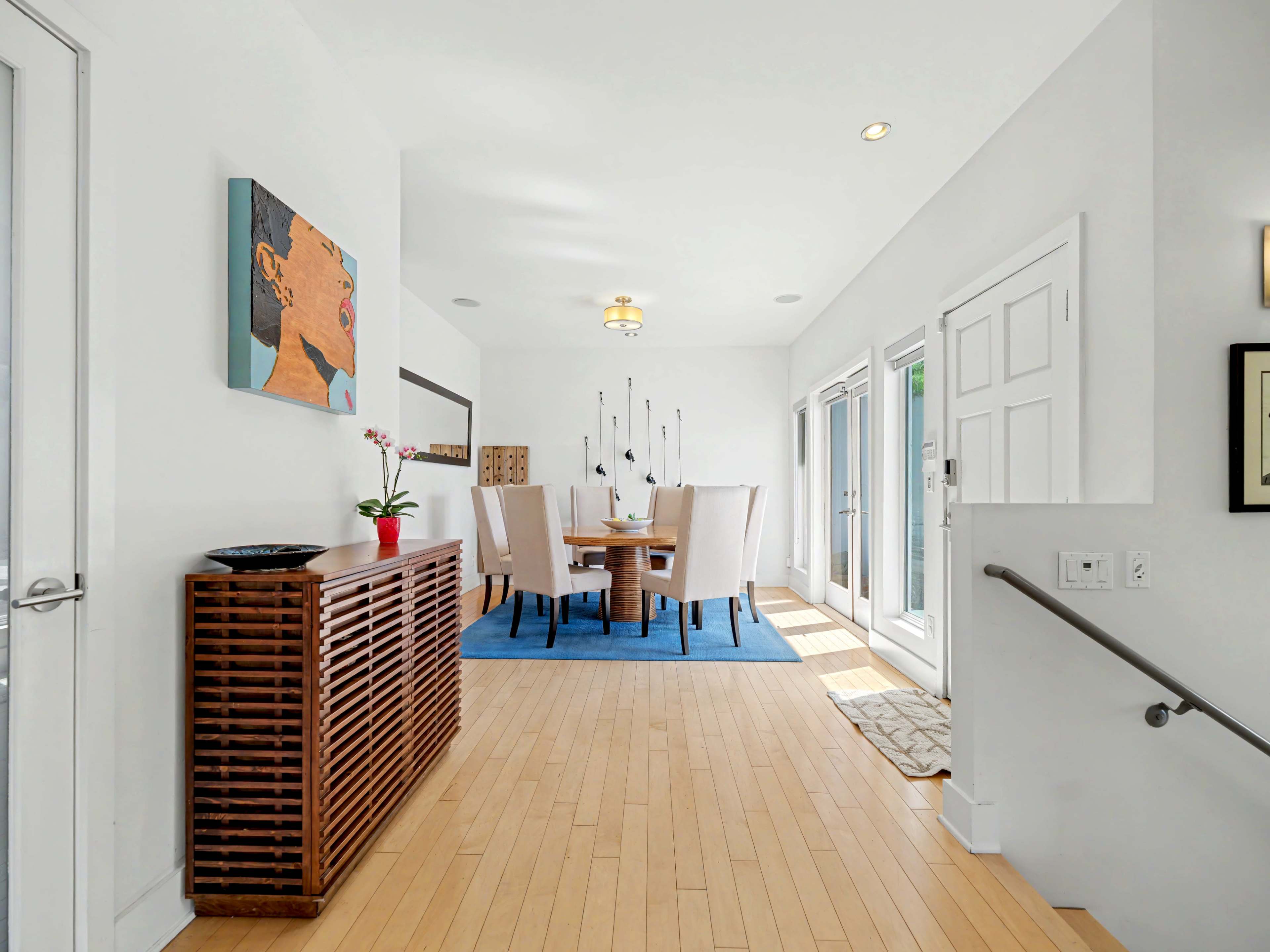 A hallway leads to a bright dining area featuring a round table with six chairs, a blue area rug, and large windows letting in natural light.