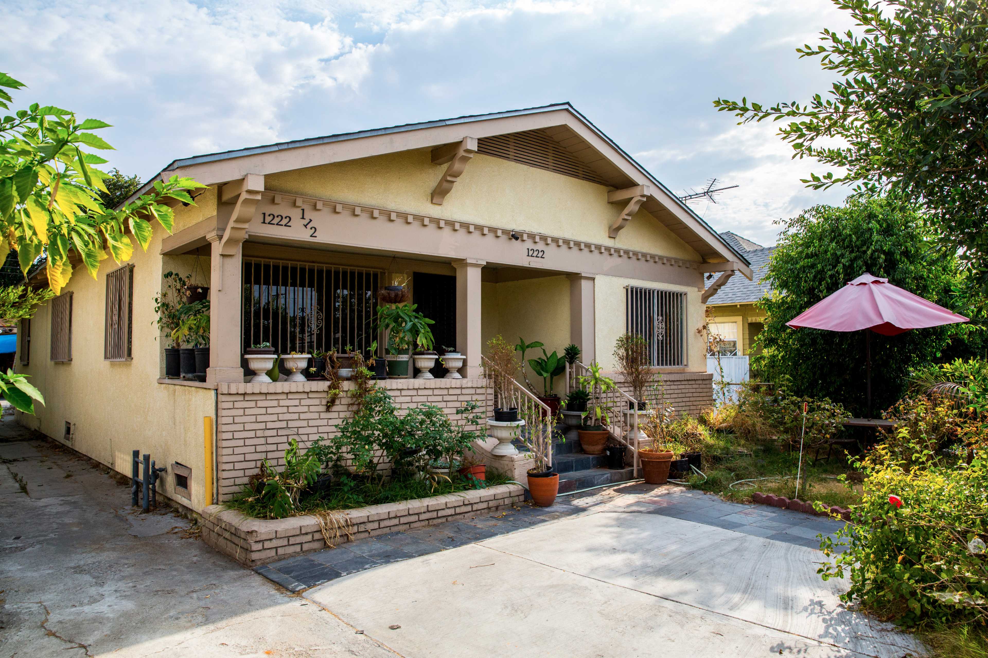 The image shows a single-story house with a covered porch, surrounded by various potted plants and trees, and a driveway leading to the front door.