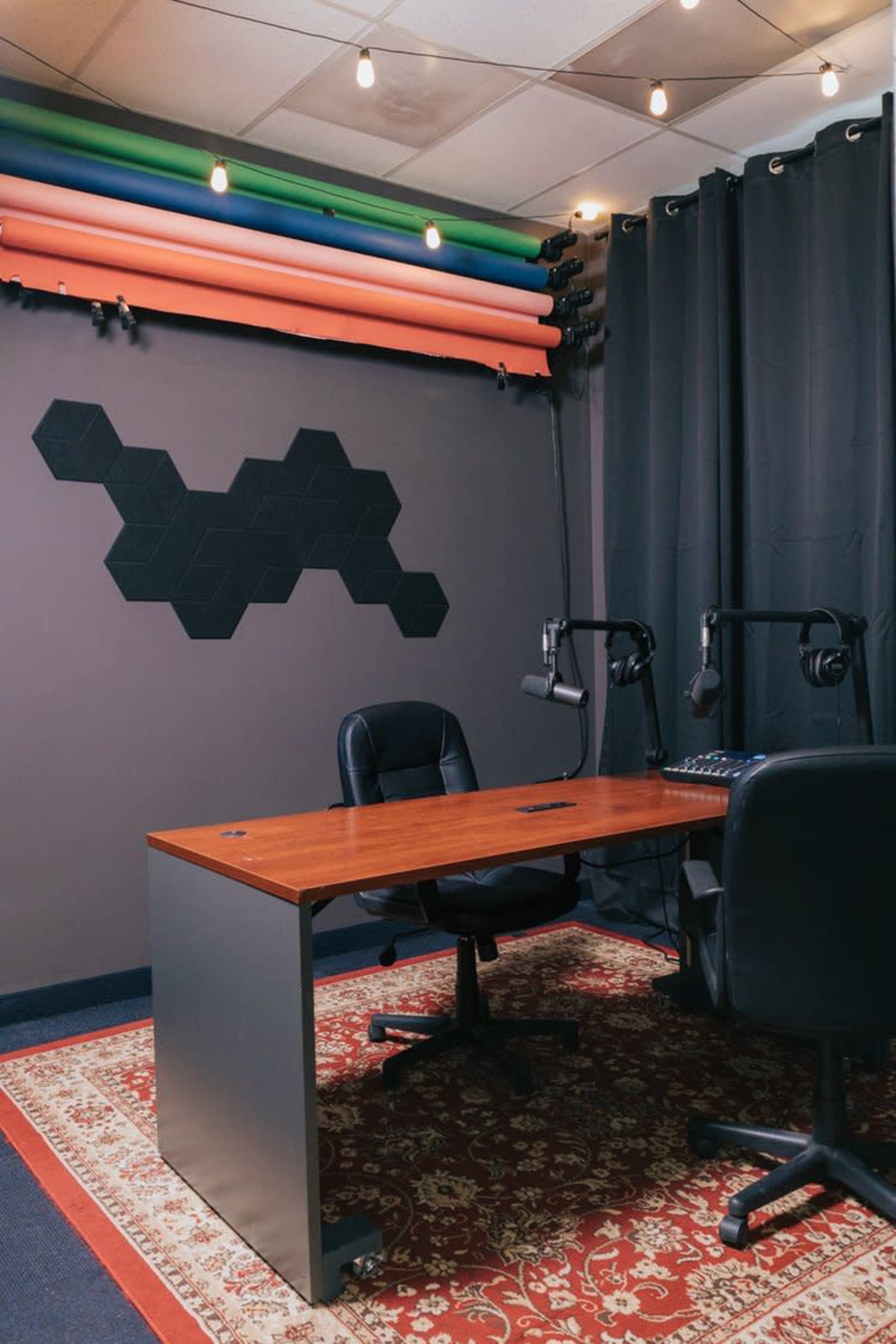 A small recording studio with a wooden table, two chairs, and a backdrop of colorful foam panels and hexagonal wall decor.