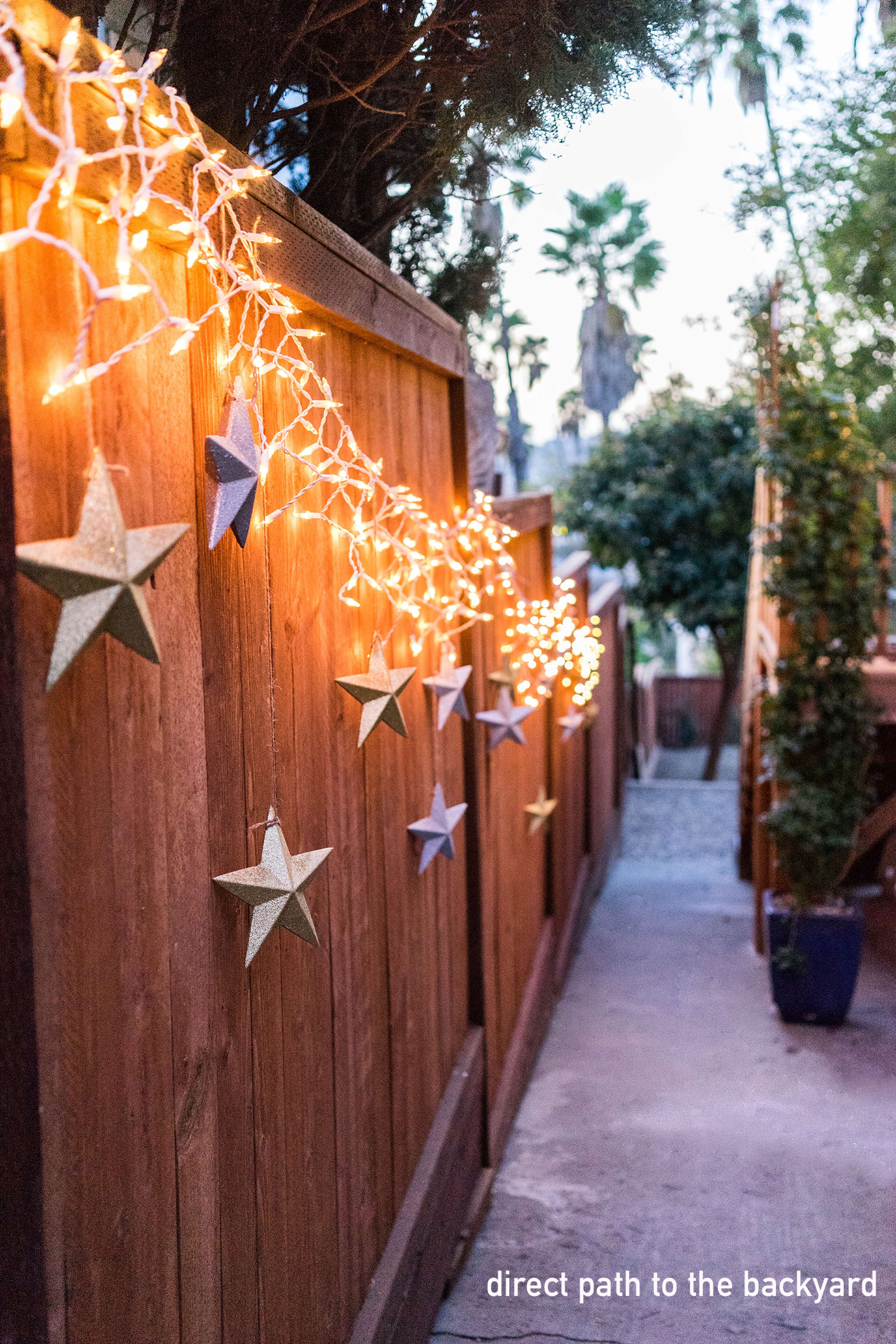 A narrow pathway bordered by a wooden fence adorned with string lights and decorative stars, leading to a backyard.