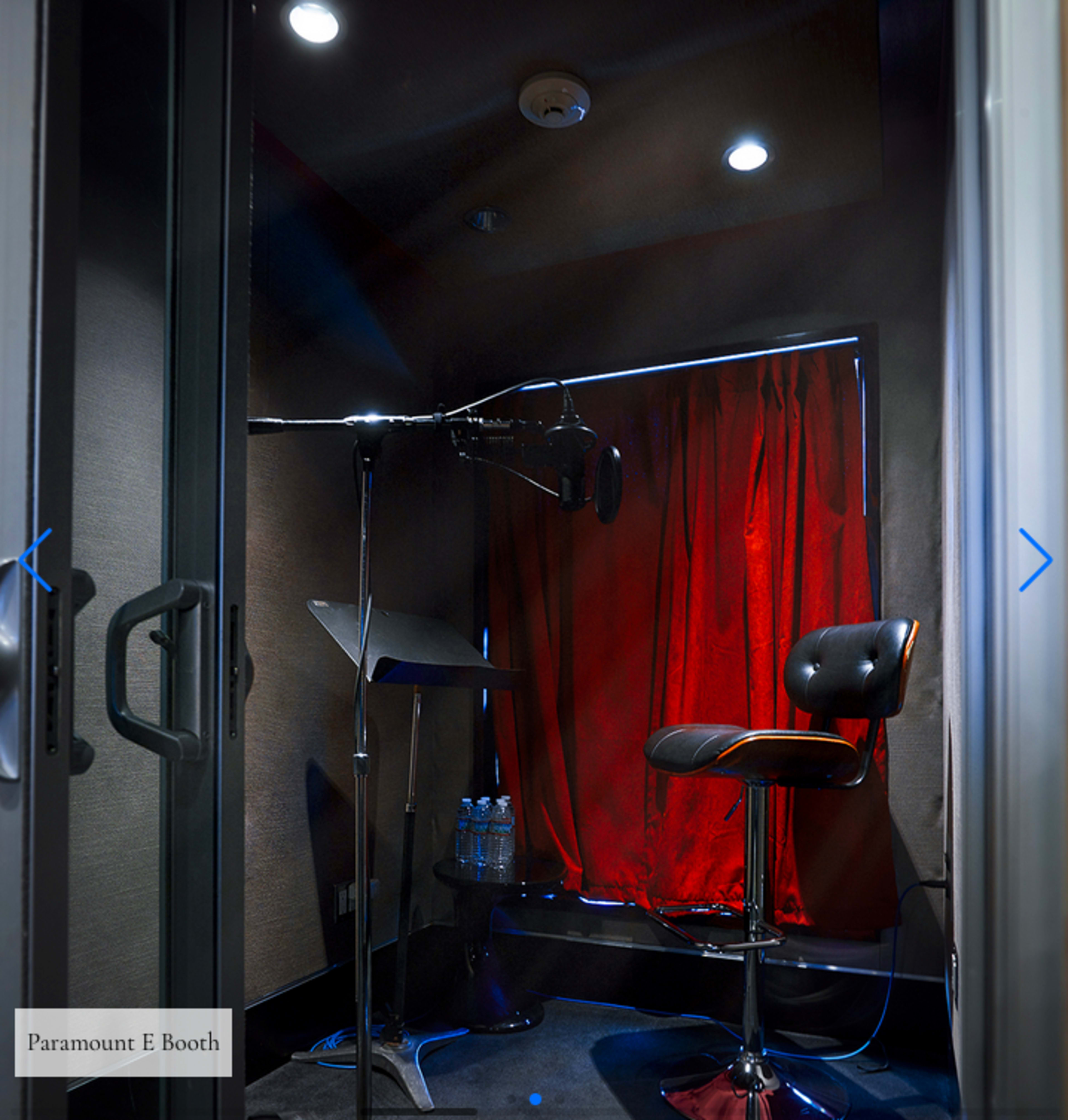 A small recording booth with a microphone, a music stand, a stool, and a red curtain.
