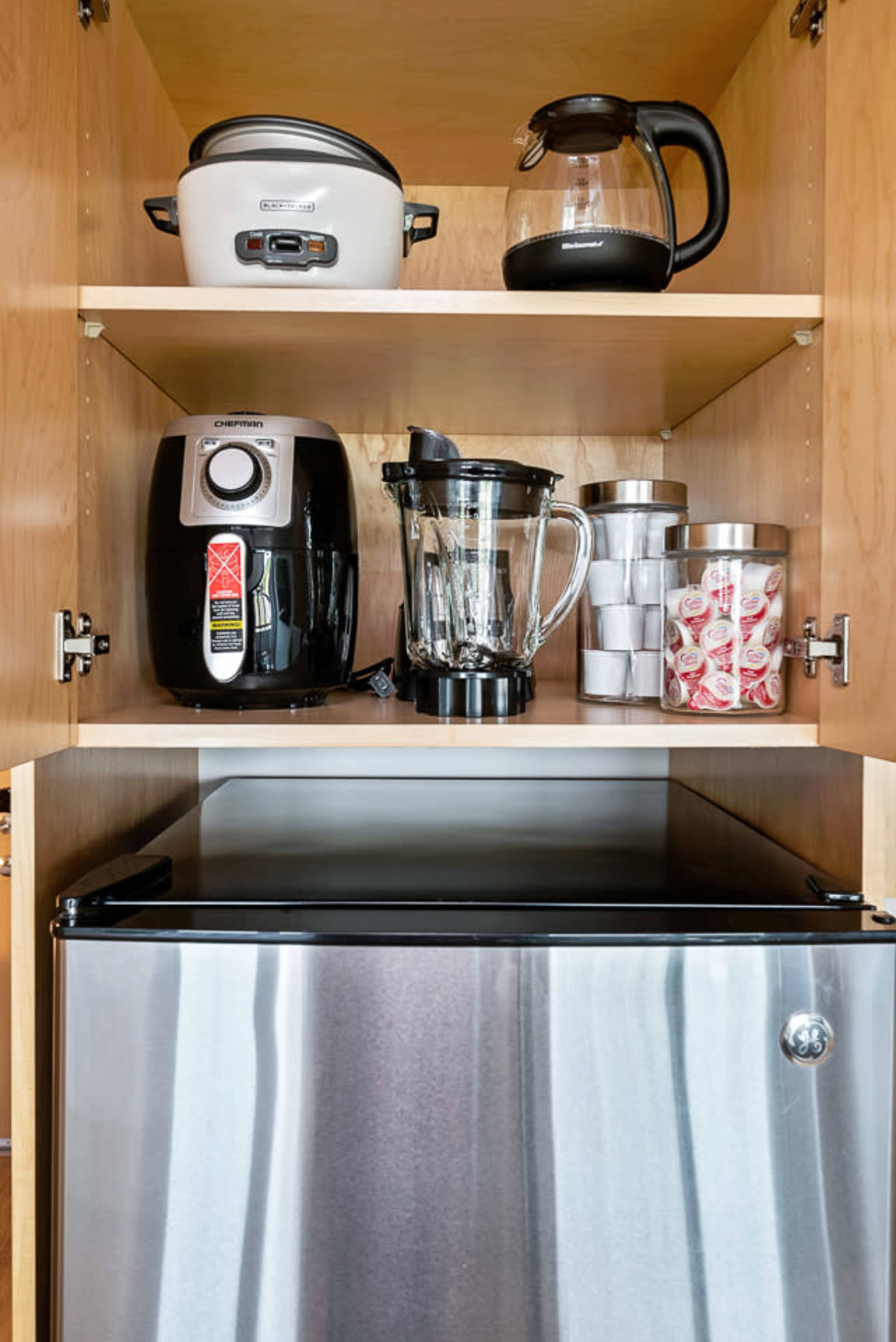 The image shows a kitchen cabinet with a rice cooker, coffee maker, blender, a container of sugar packets, and a mini fridge.