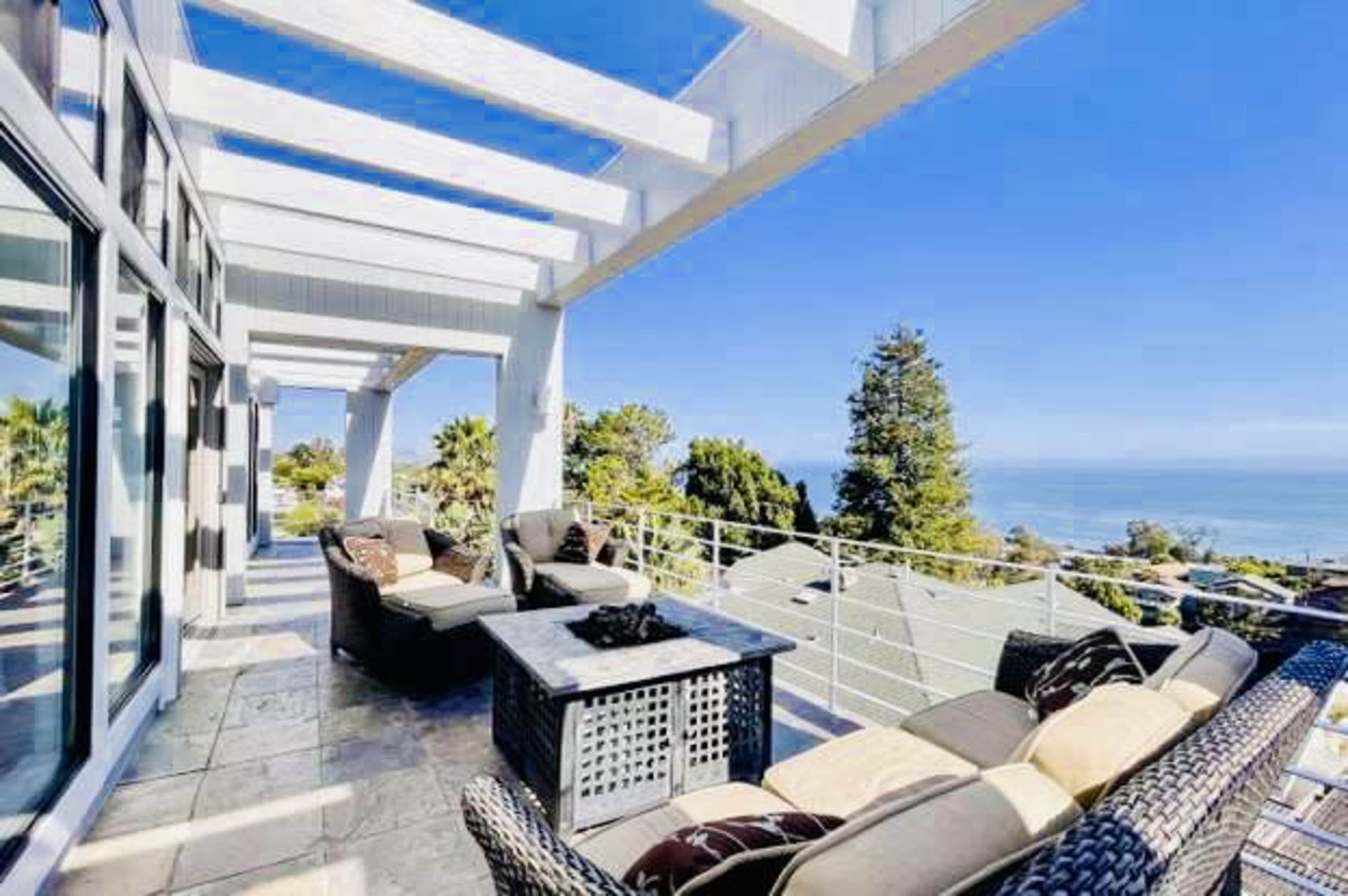 A spacious outdoor patio features seating arrangements with a fire pit, overlooking a coastal view under a clear blue sky.