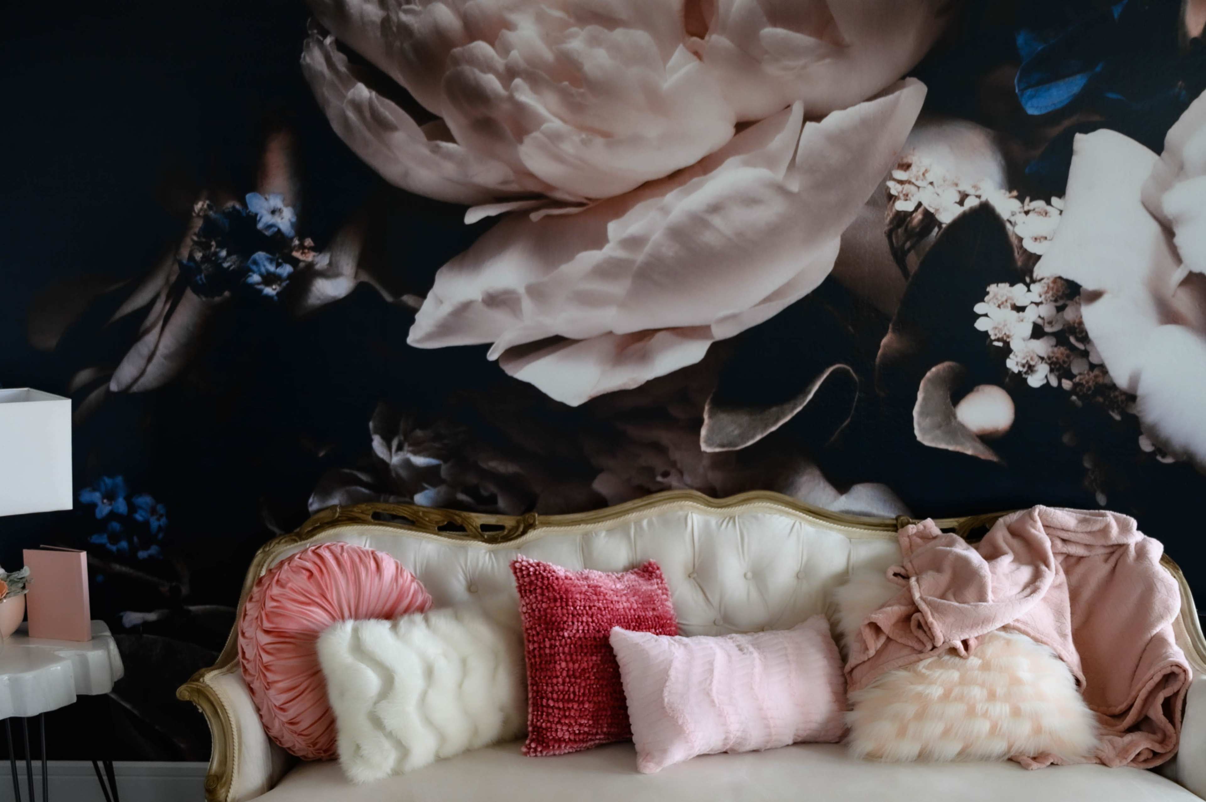 A white vintage sofa with various pink and white cushions is positioned against a floral backdrop featuring large, soft-colored flowers.