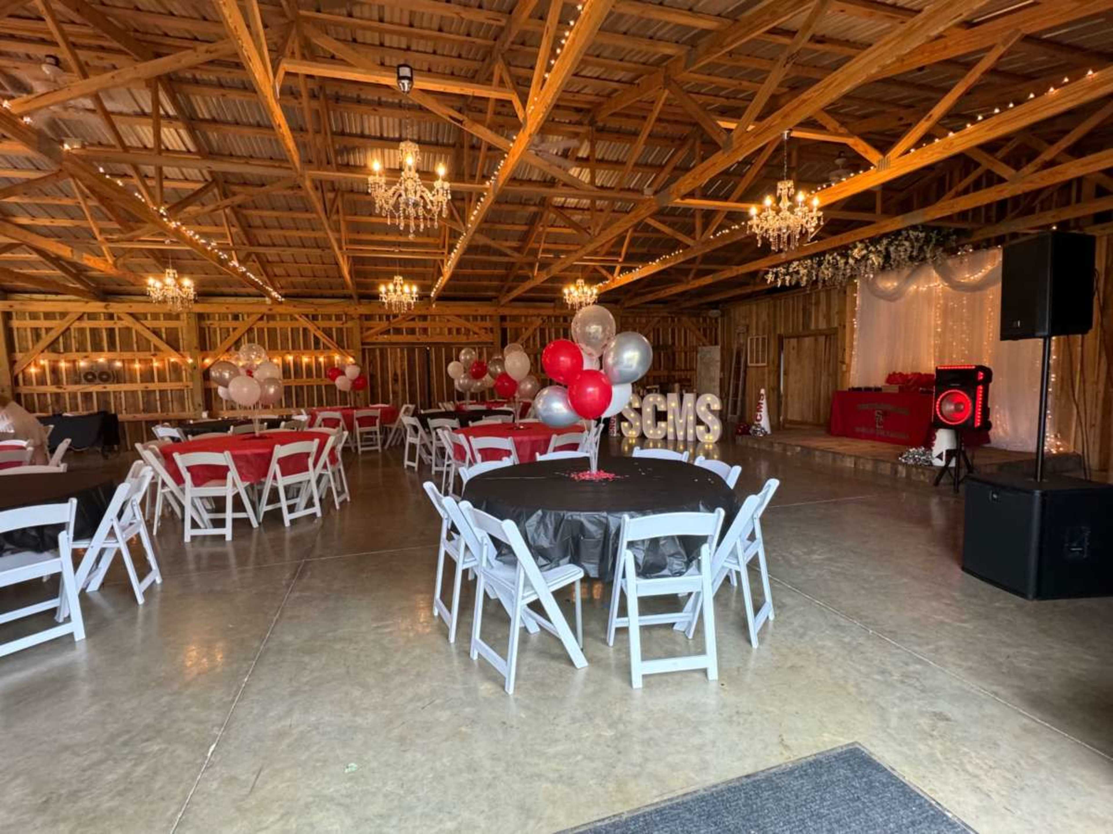 The interior of a barn is set up for an event, featuring tables with black and red tablecloths, balloons, chandeliers, and a decorated stage area.