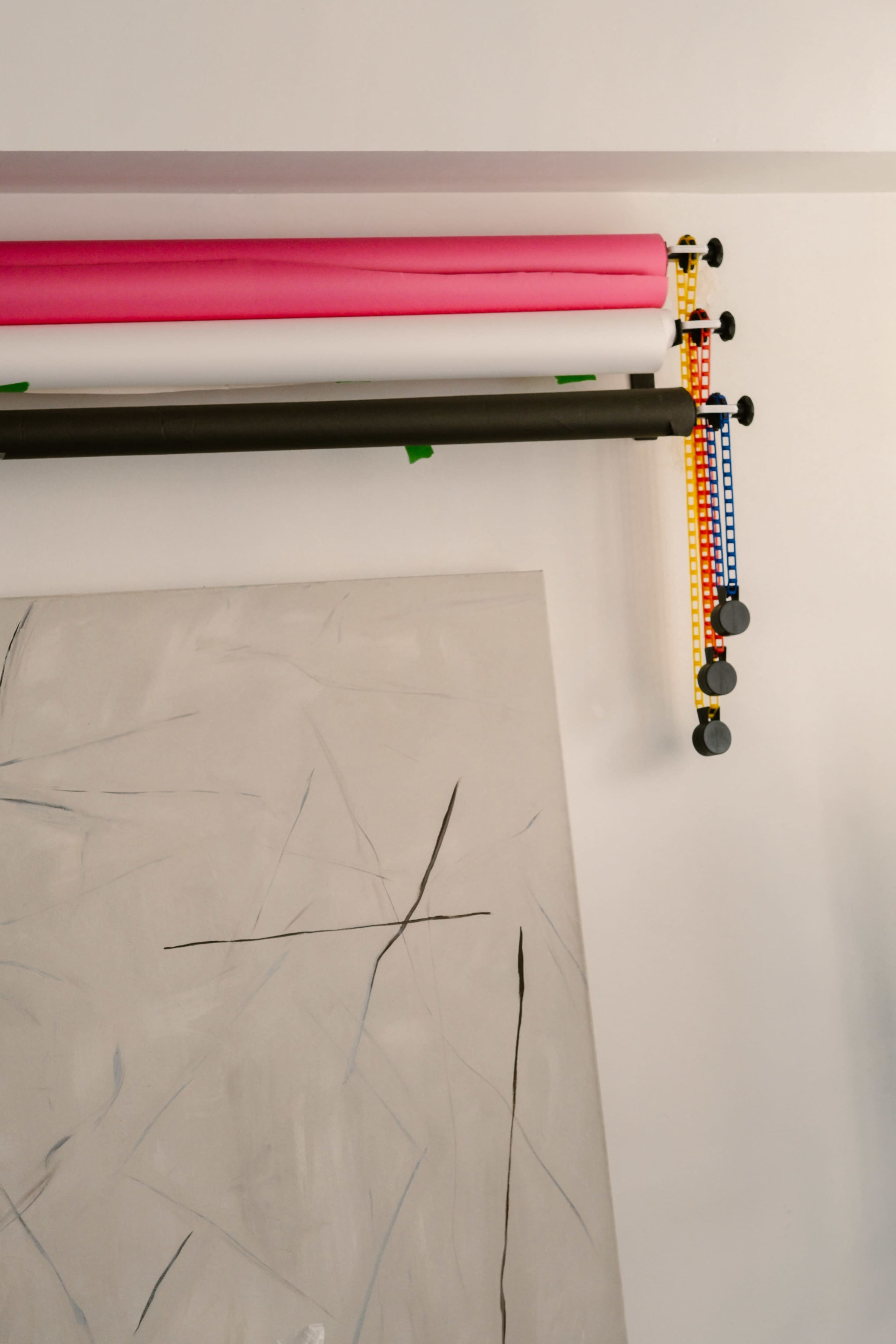 The image shows rolls of colored paper hanging on a wall above a large, textured canvas with abstract black lines.