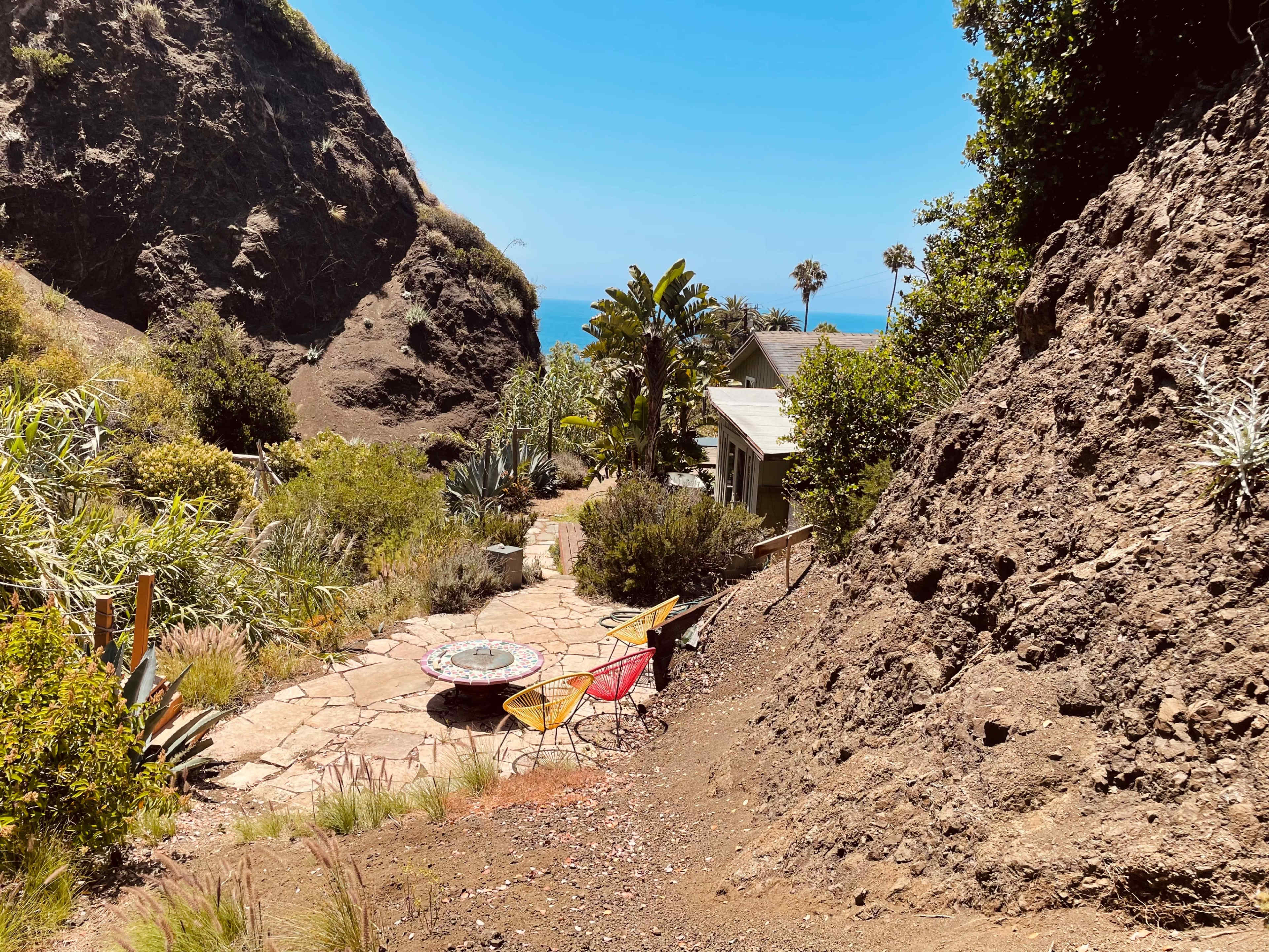 A pathway leads down through rocky terrain to a patio area with colorful chairs overlooking the ocean.