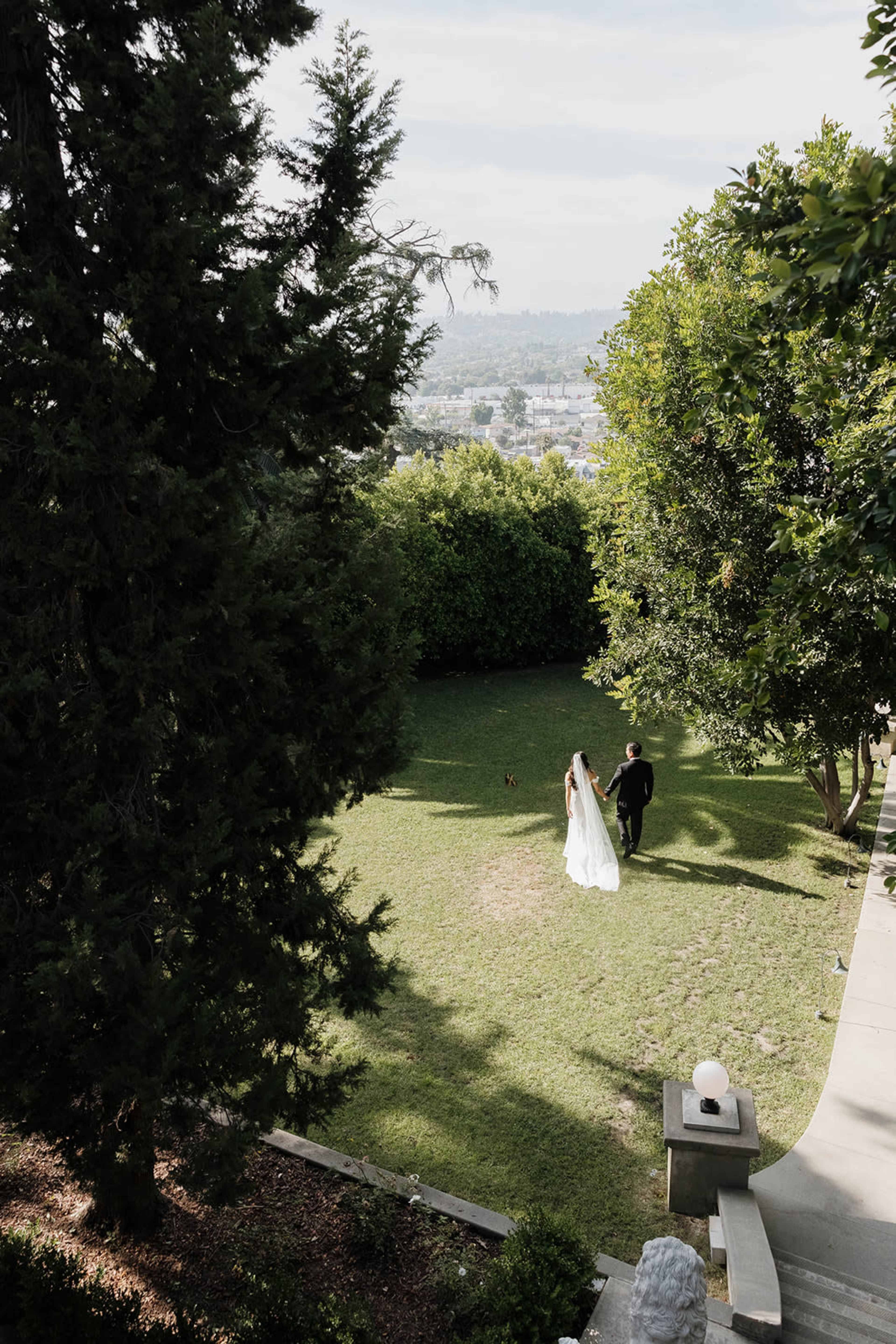 A couple walks hand-in-hand across a landscaped yard with lush green grass and trees, overlooking a distant cityscape.