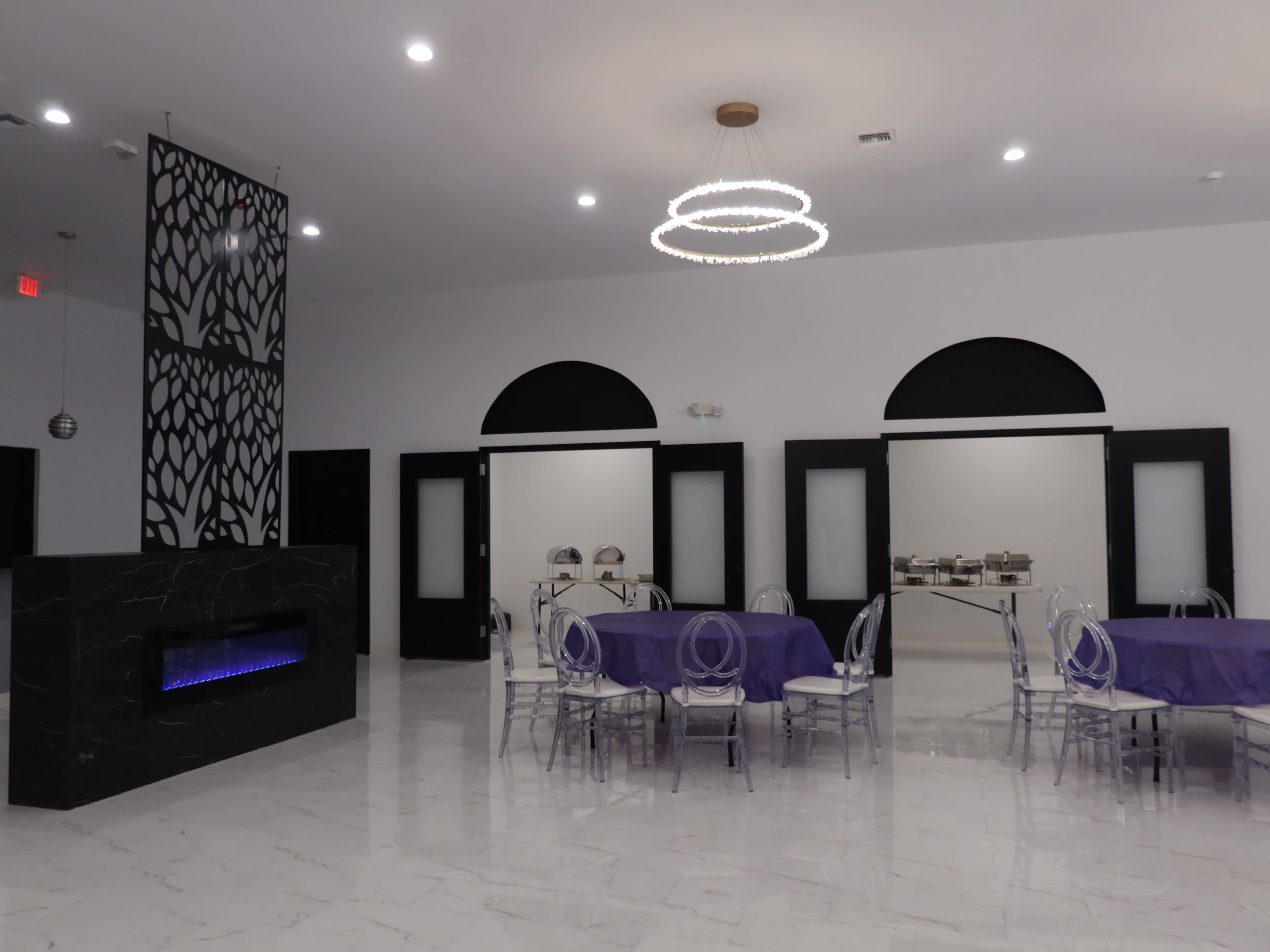 An elegant event space featuring purple tablecloths on round tables, clear acrylic chairs, a modern fireplace, and a decorative wall with a black design.