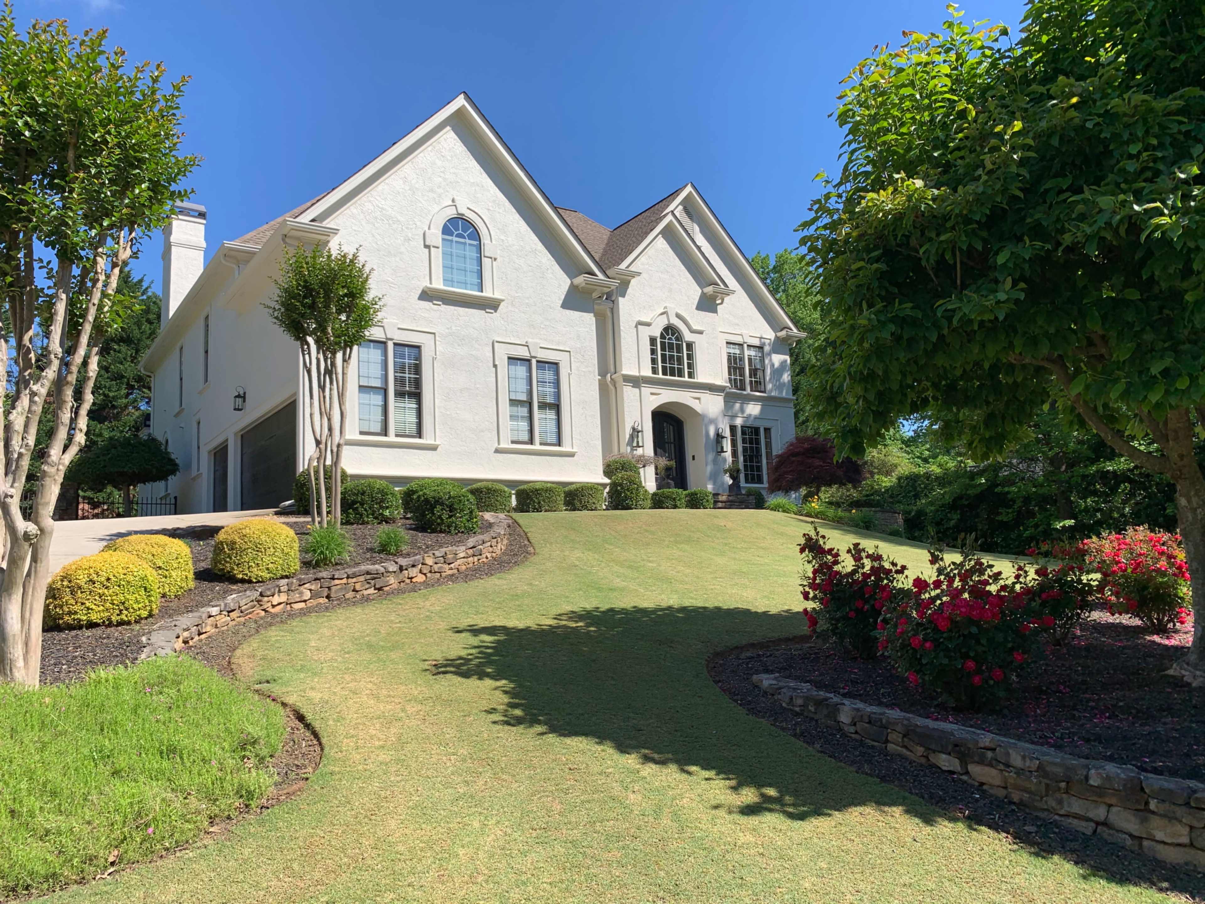 A large white house with a steeply pitched roof is set on a well-manicured lawn surrounded by neatly trimmed shrubs and flower beds.
