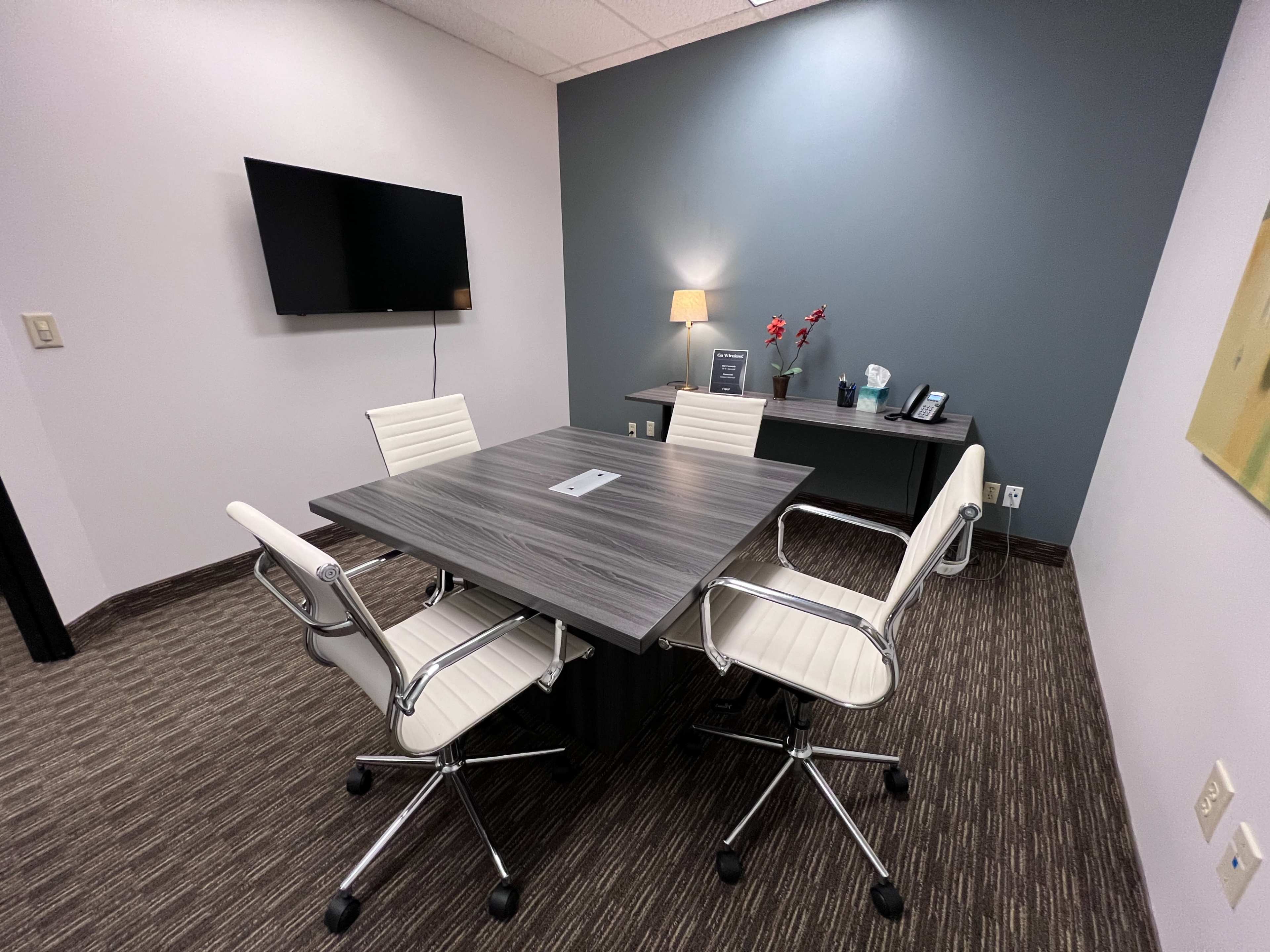 A modern conference room features a square table with four white chairs, a wall-mounted television, and a desk against a gray and white backdrop.