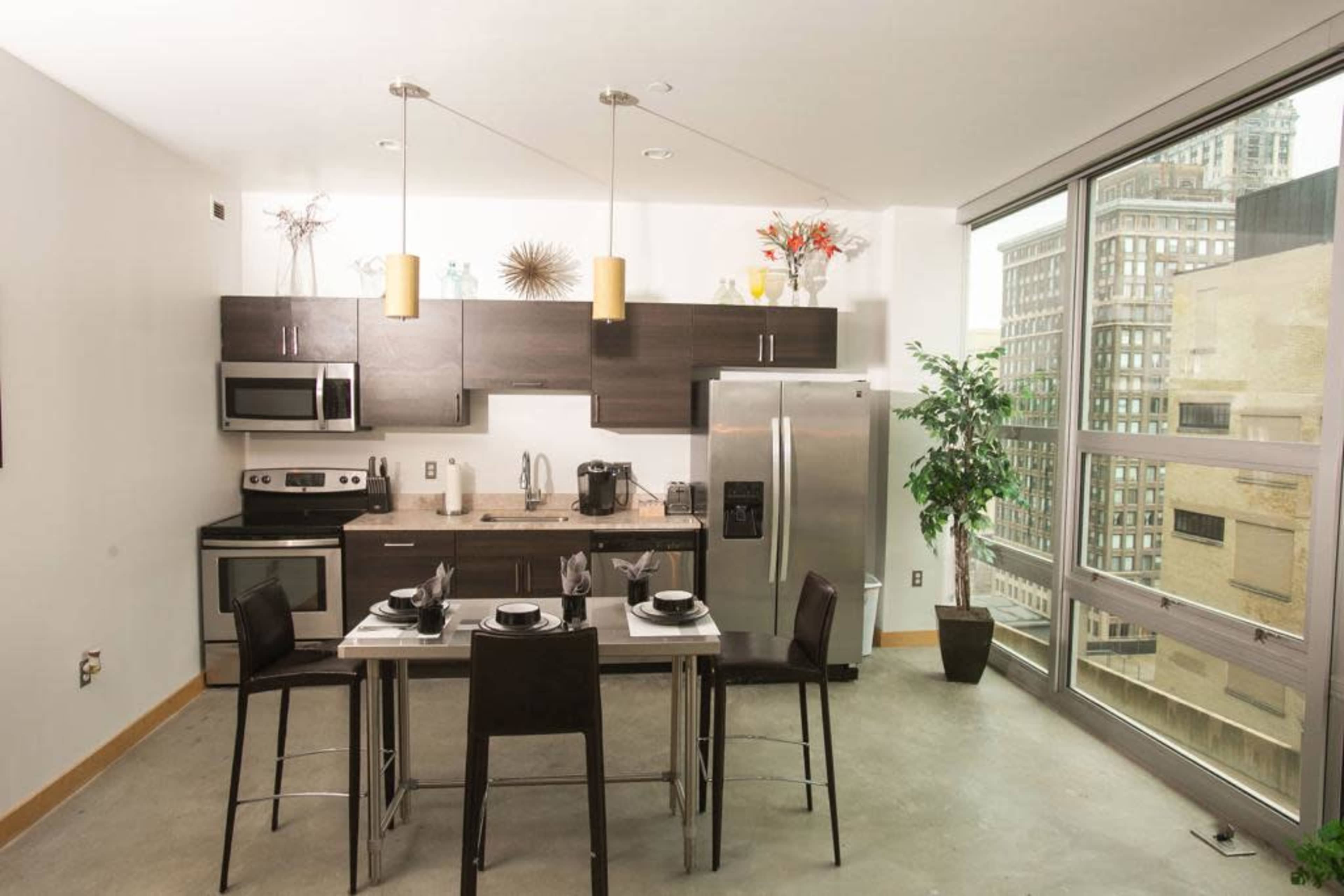 A modern kitchen features stainless steel appliances, dark cabinetry, a dining table set for four, and large windows overlooking a cityscape.