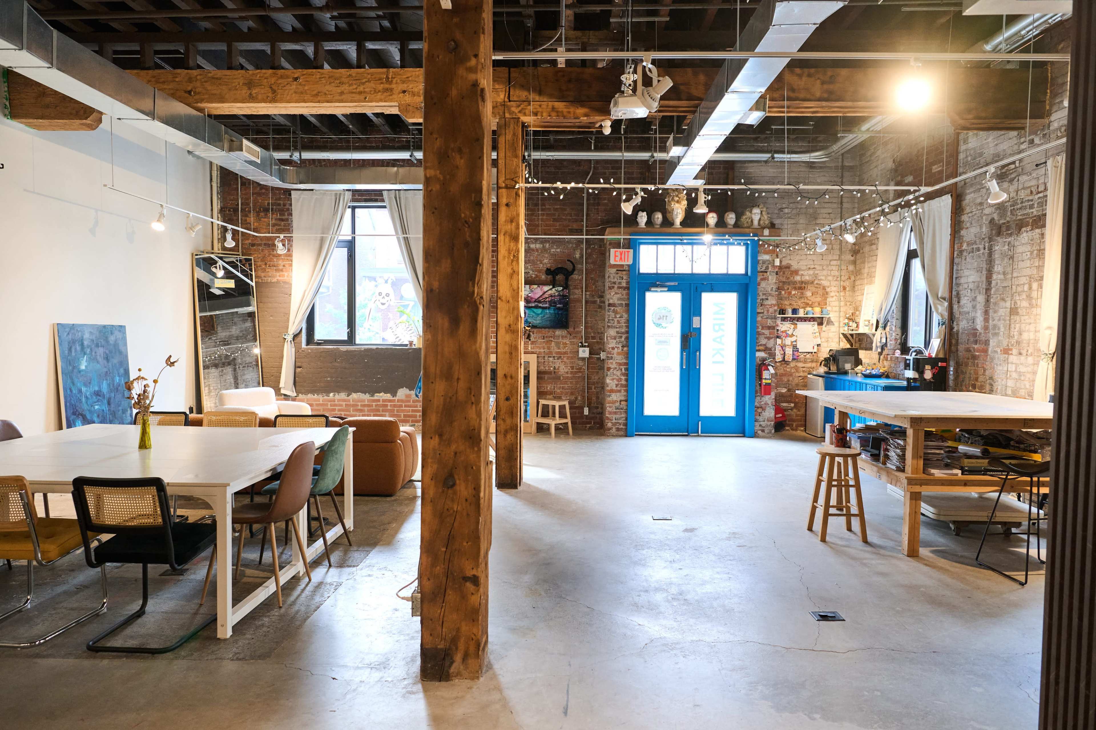 The image shows a spacious, industrial-style room with exposed brick walls, wooden beams, a large table, and a blue door leading outside.