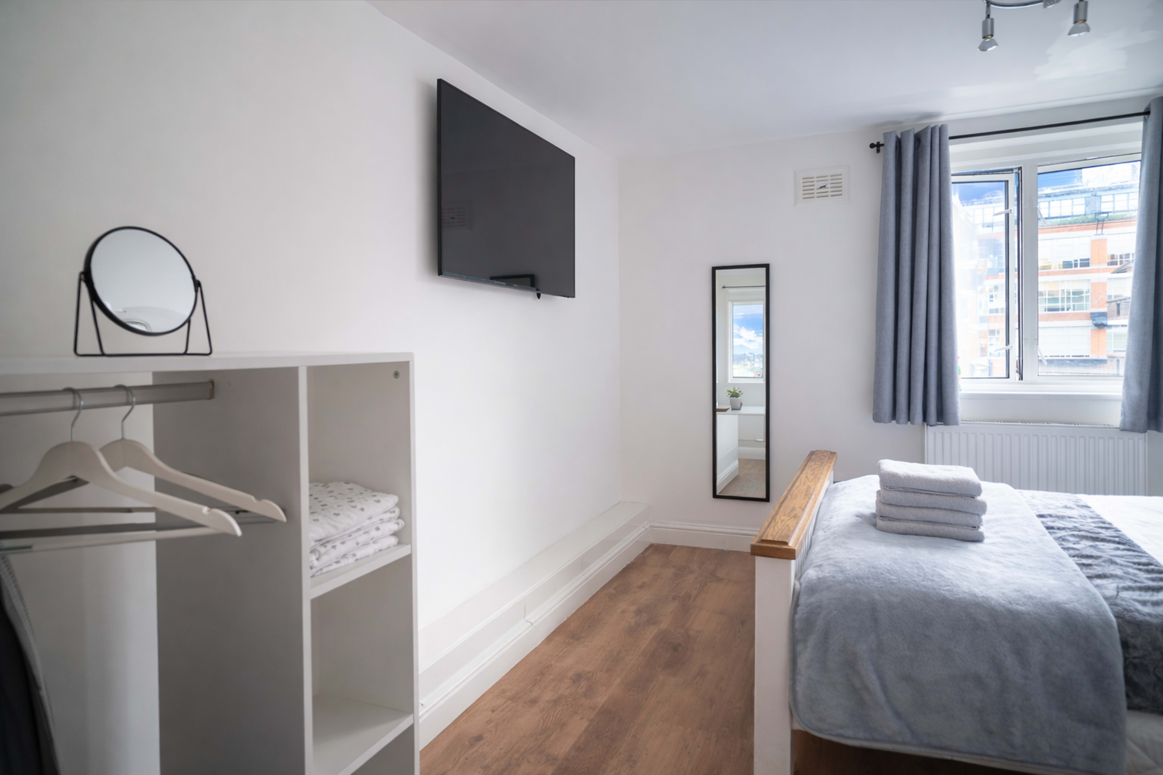 The image shows a modern bedroom featuring a wall-mounted television, a large mirror, a small storage unit, and neatly arranged bedding with towels on the bed.