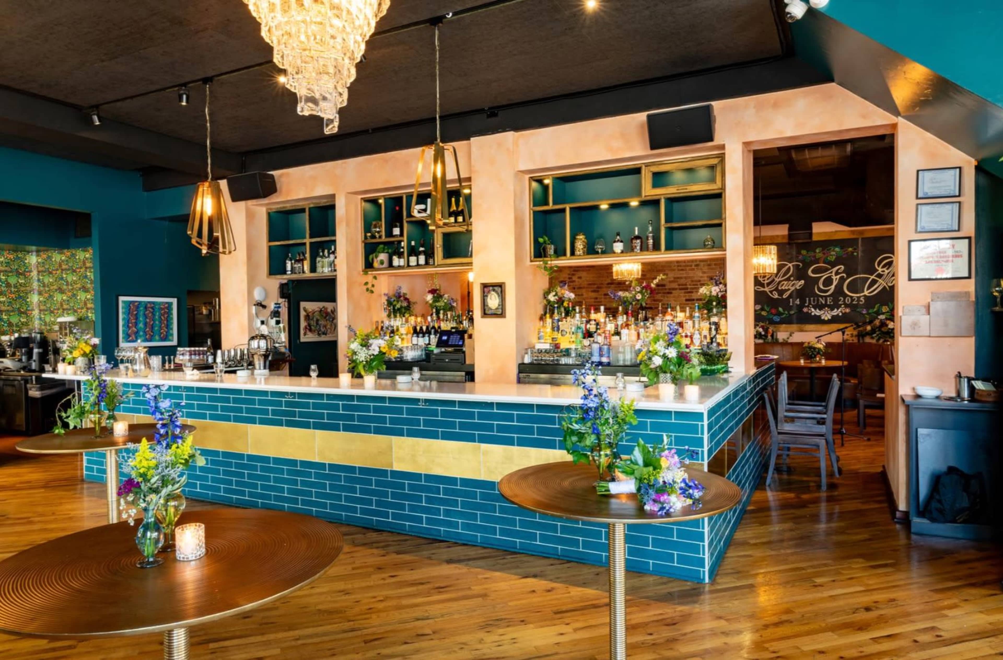 The image shows a vibrant bar area with a blue tile front, a chandelier overhead, and tables adorned with flower centerpieces.