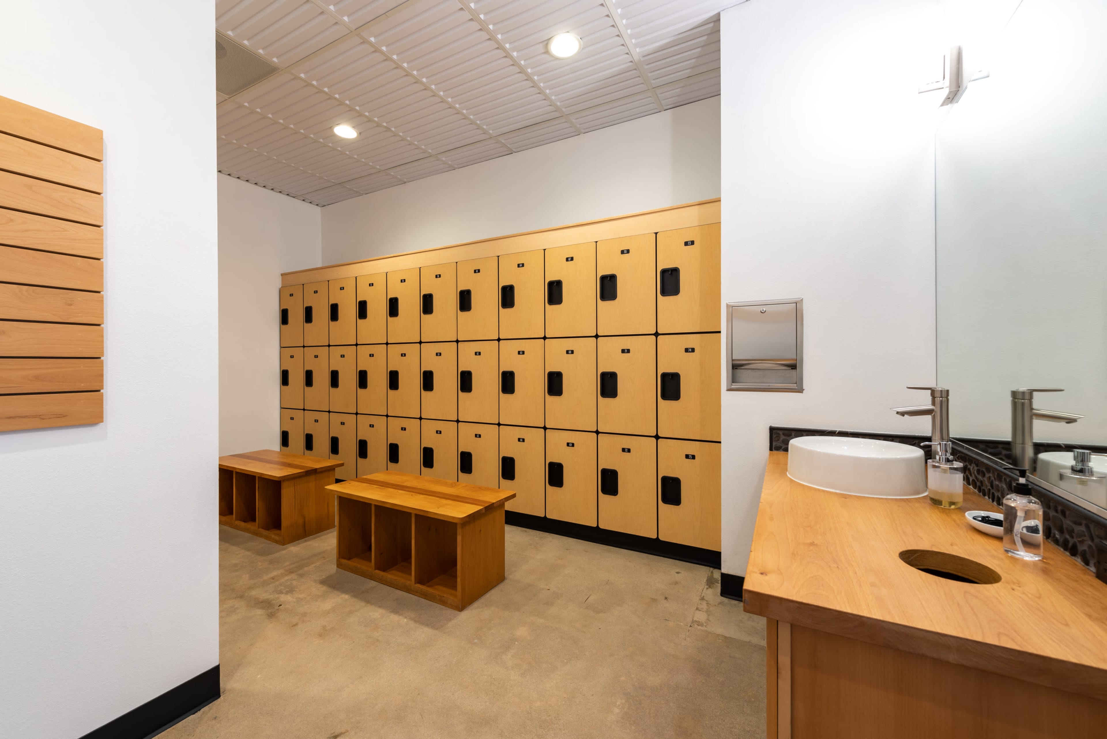 The image shows a locker room with wooden lockers lining one wall, two benches in front, and a sink area on the opposite wall.