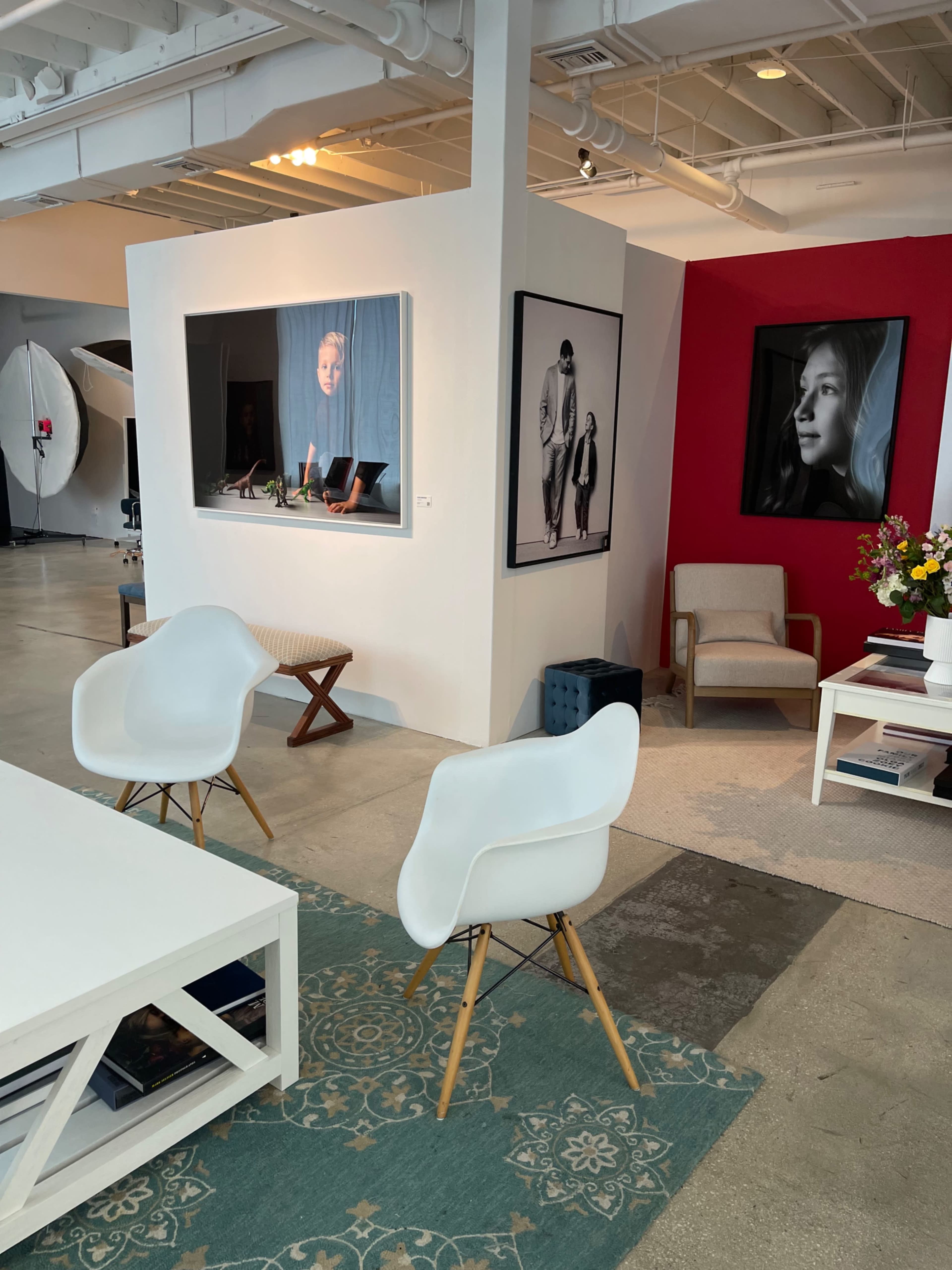 The image shows a modern art gallery space with white walls, a red accent wall featuring black-and-white photographs, and minimalist furniture.