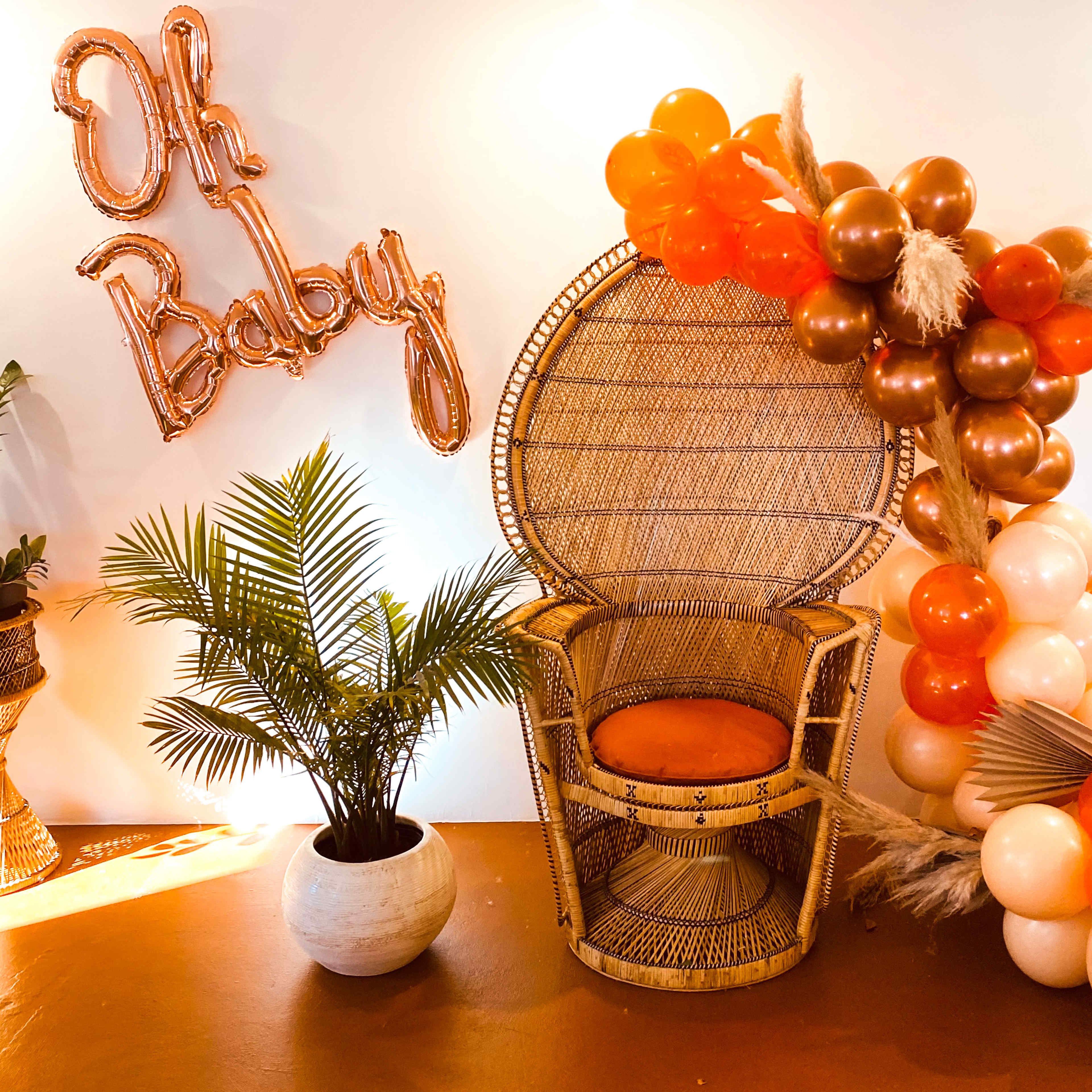 A decorative setup featuring a woven chair with an orange cushion, surrounded by a plant, and adorned with an "Oh Baby" balloon banner and colorful balloons.