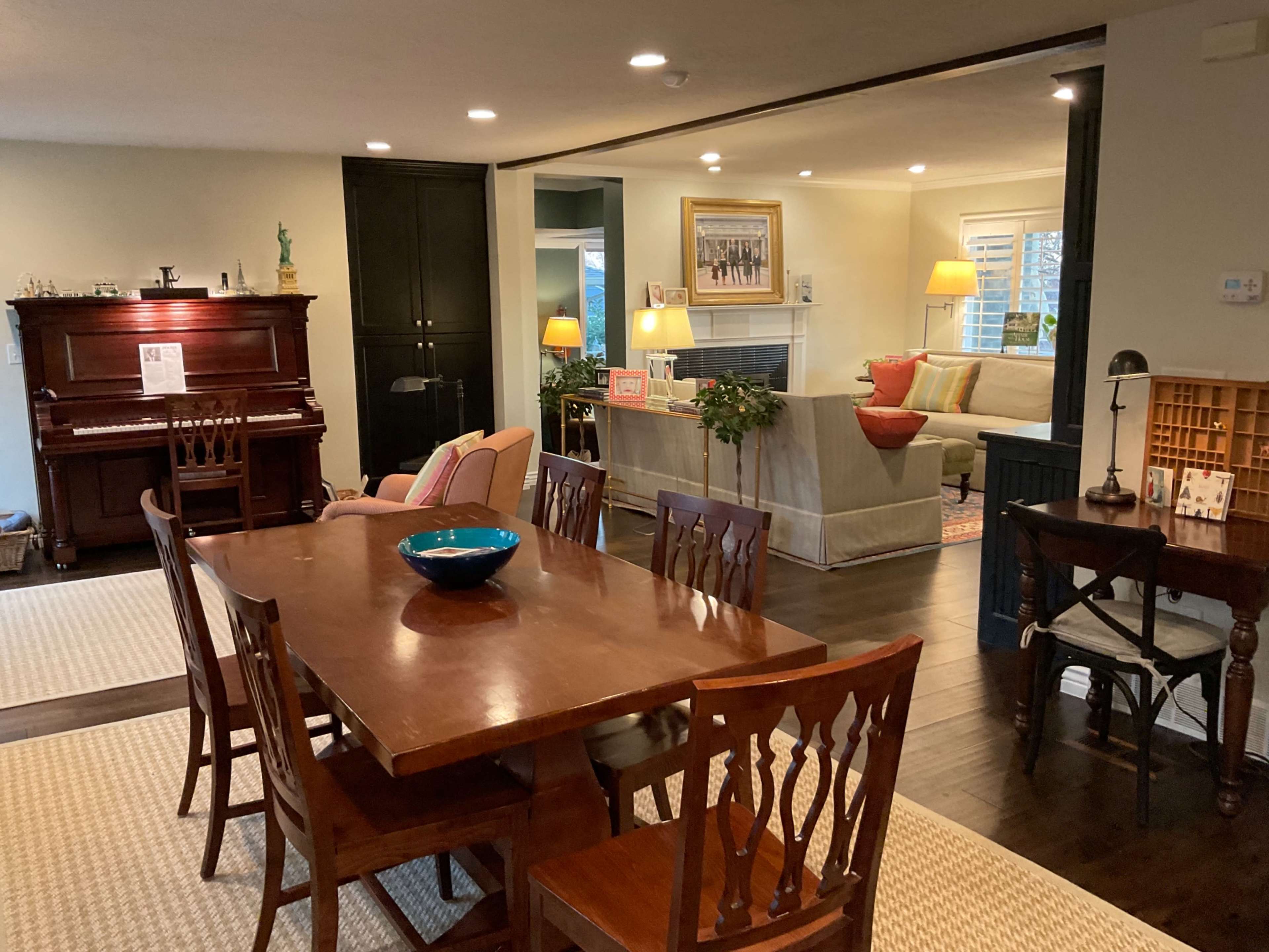 A dining area with a wooden table and chairs is situated near a living room that features a piano, plants, and soft lighting.