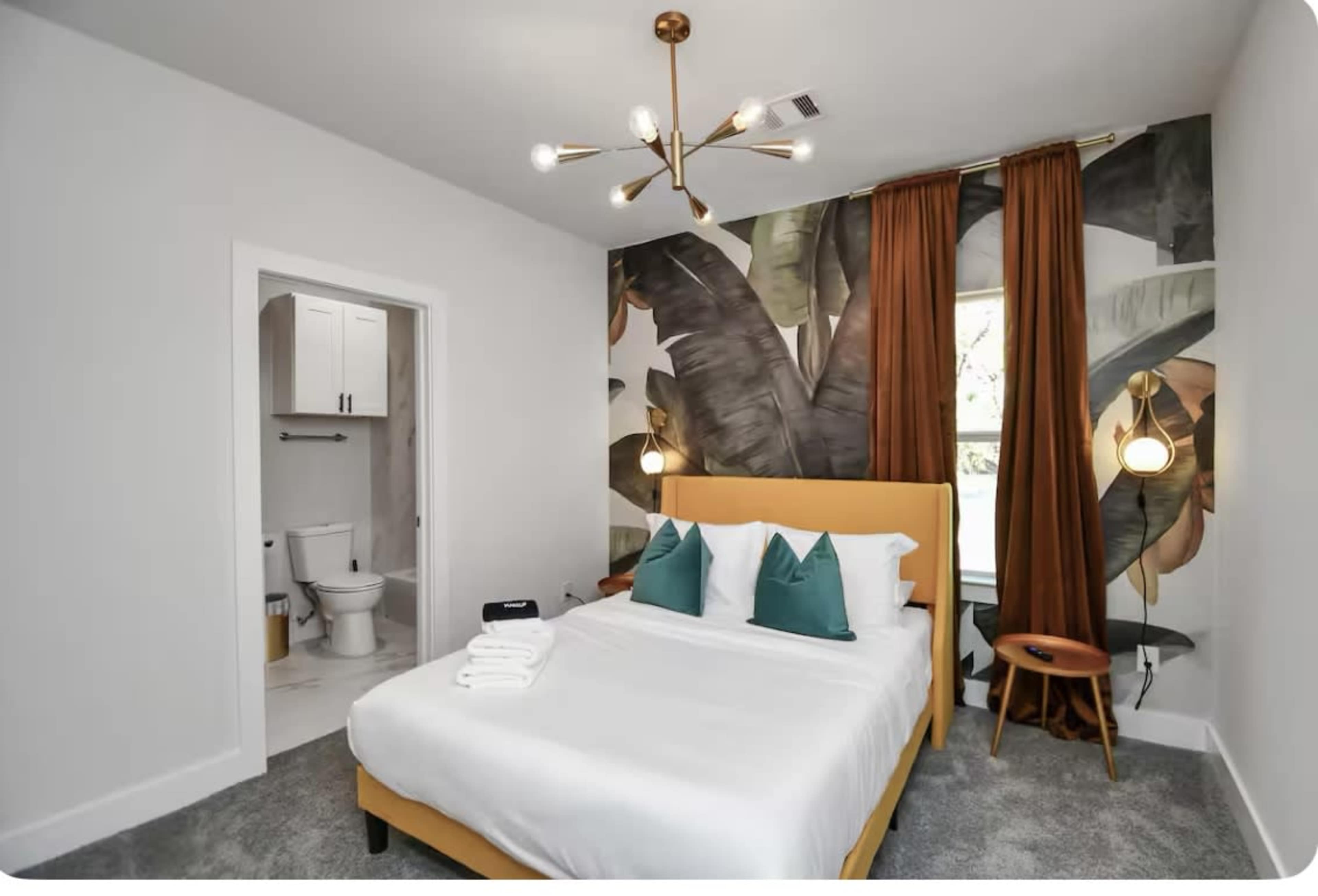 The image shows a well-decorated bedroom featuring a yellow bed frame, two green cushions, and a noticeable leaf-patterned wallpaper, with a bathroom visible in the background.