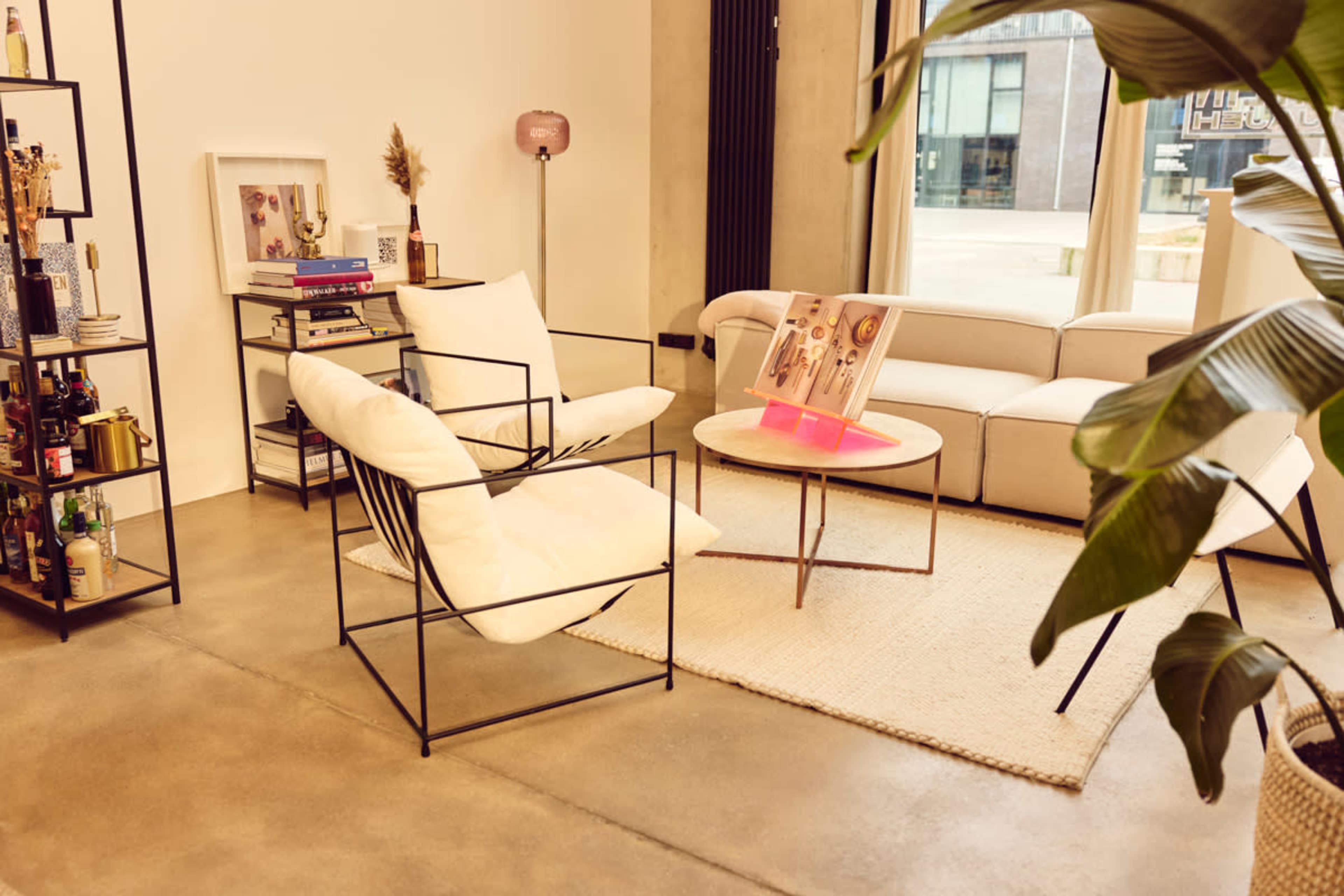 The image shows a modern living room with a light-colored sofa, a round coffee table, two stylish chairs, and decorative elements on a media console.
