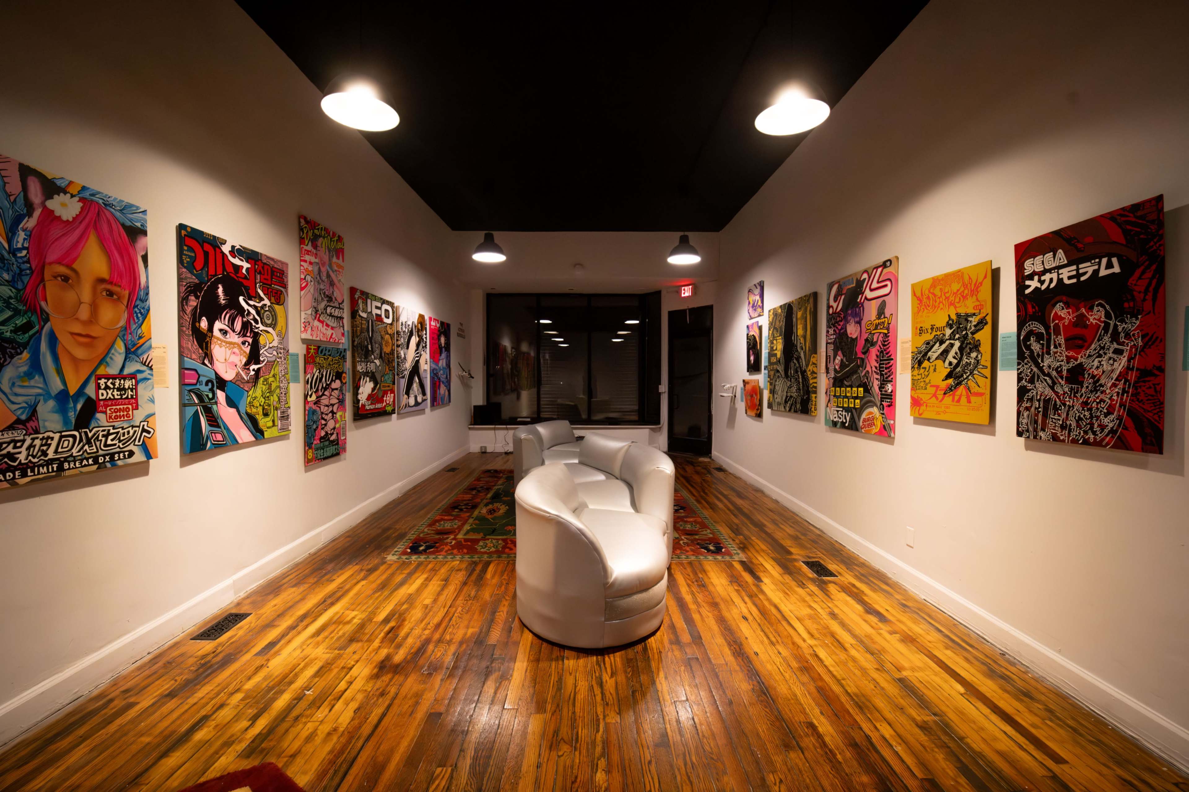 The image shows a narrow art gallery with white walls adorned with colorful posters, featuring a minimalist seating arrangement in the center and dark wooden flooring.