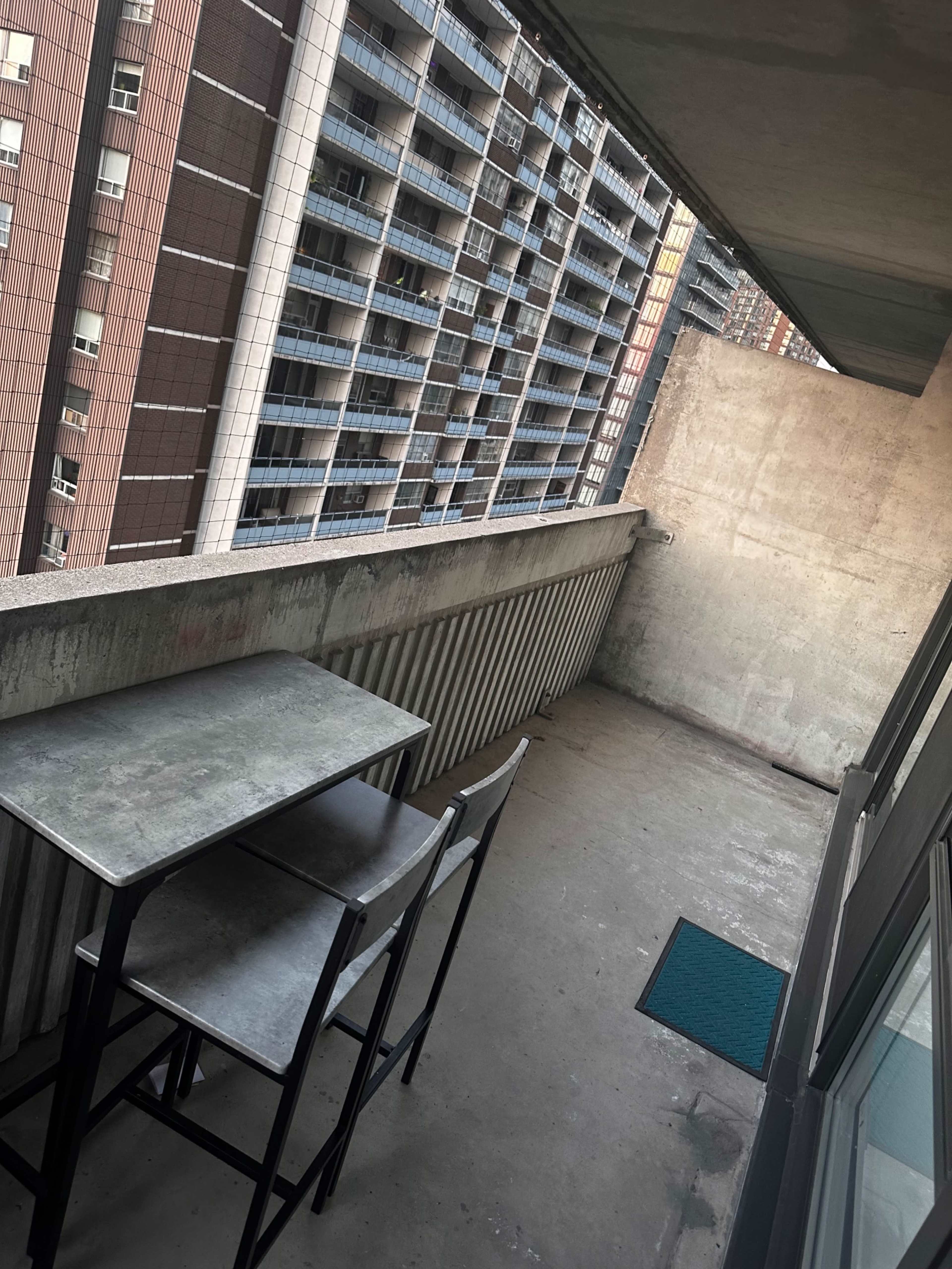 The image shows a balcony with a metal table and two chairs facing a concrete wall, overlooking a view of tall buildings.