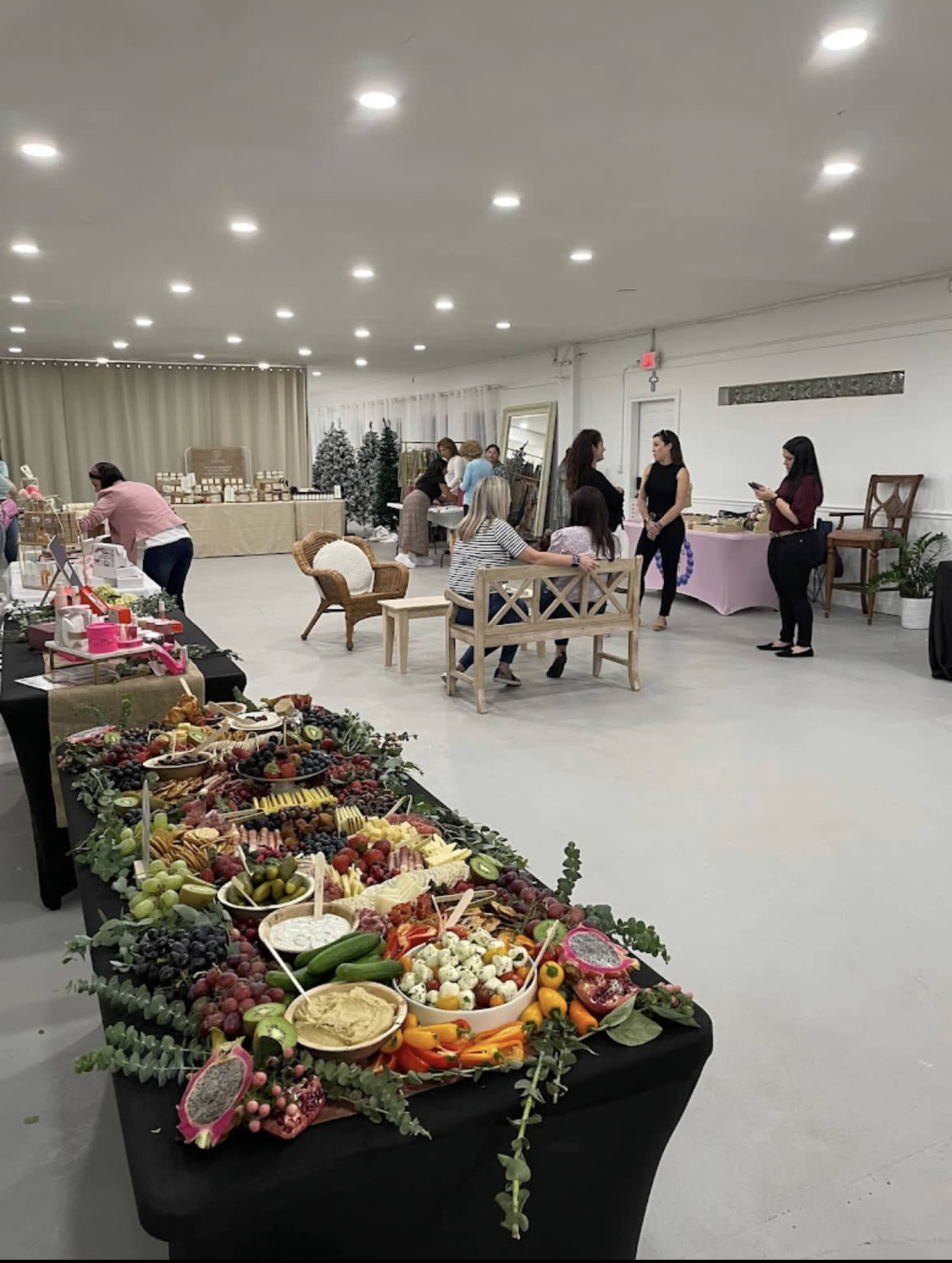 A event space is set up with a large display of assorted fruits and snacks on a black table, while people mingle in the background.