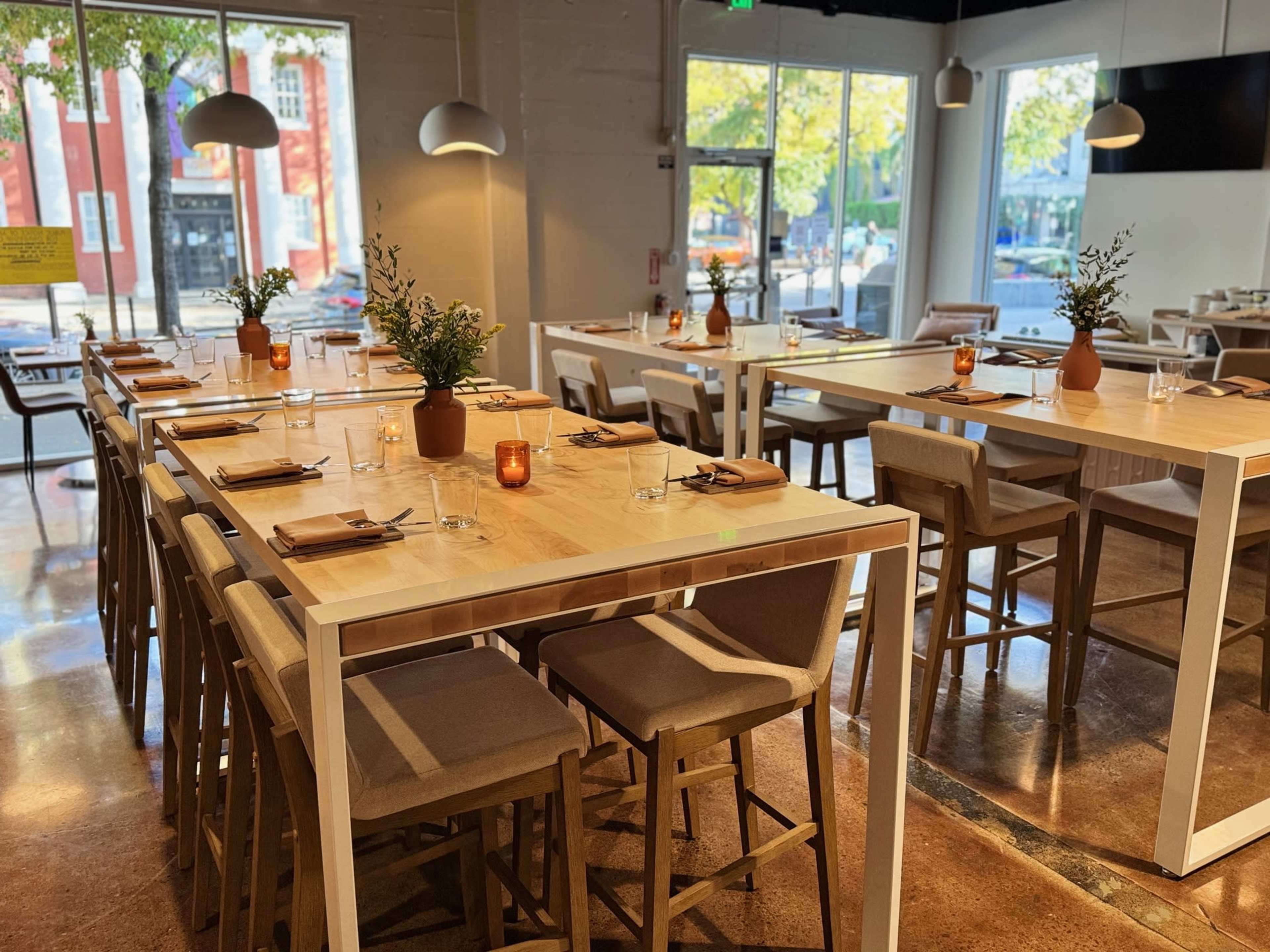The image shows a modern dining area with wooden tables, high chairs, and decorative plants on each table, illuminated by large pendant lights.