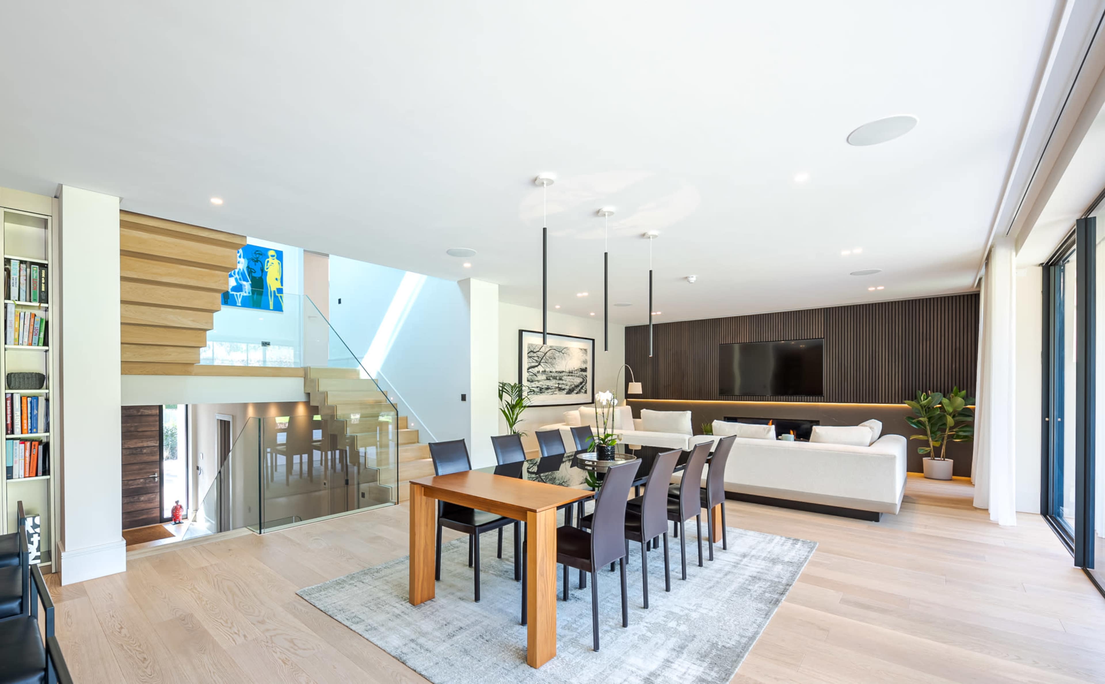 A spacious living and dining area features a wooden dining table with chairs, a staircase, and a large seating area with a television on the wall.
