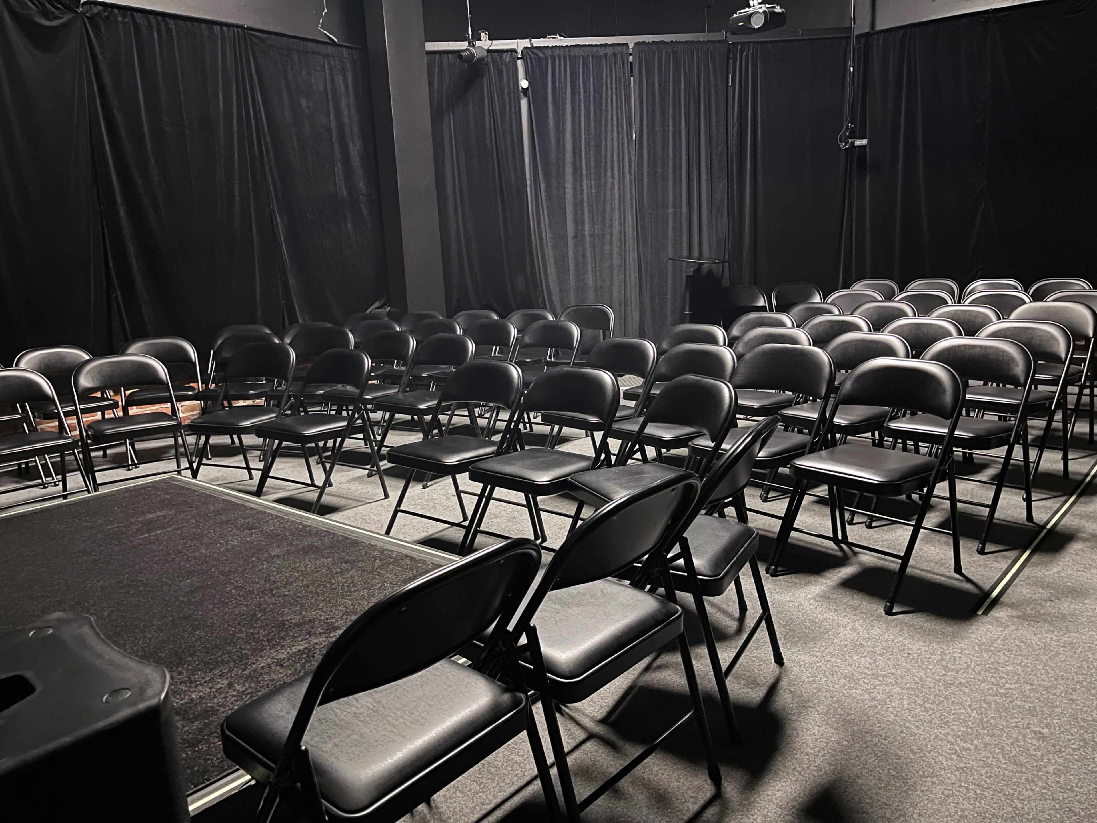 A row of black folding chairs is arranged in front of a small stage in a dimly lit theater.