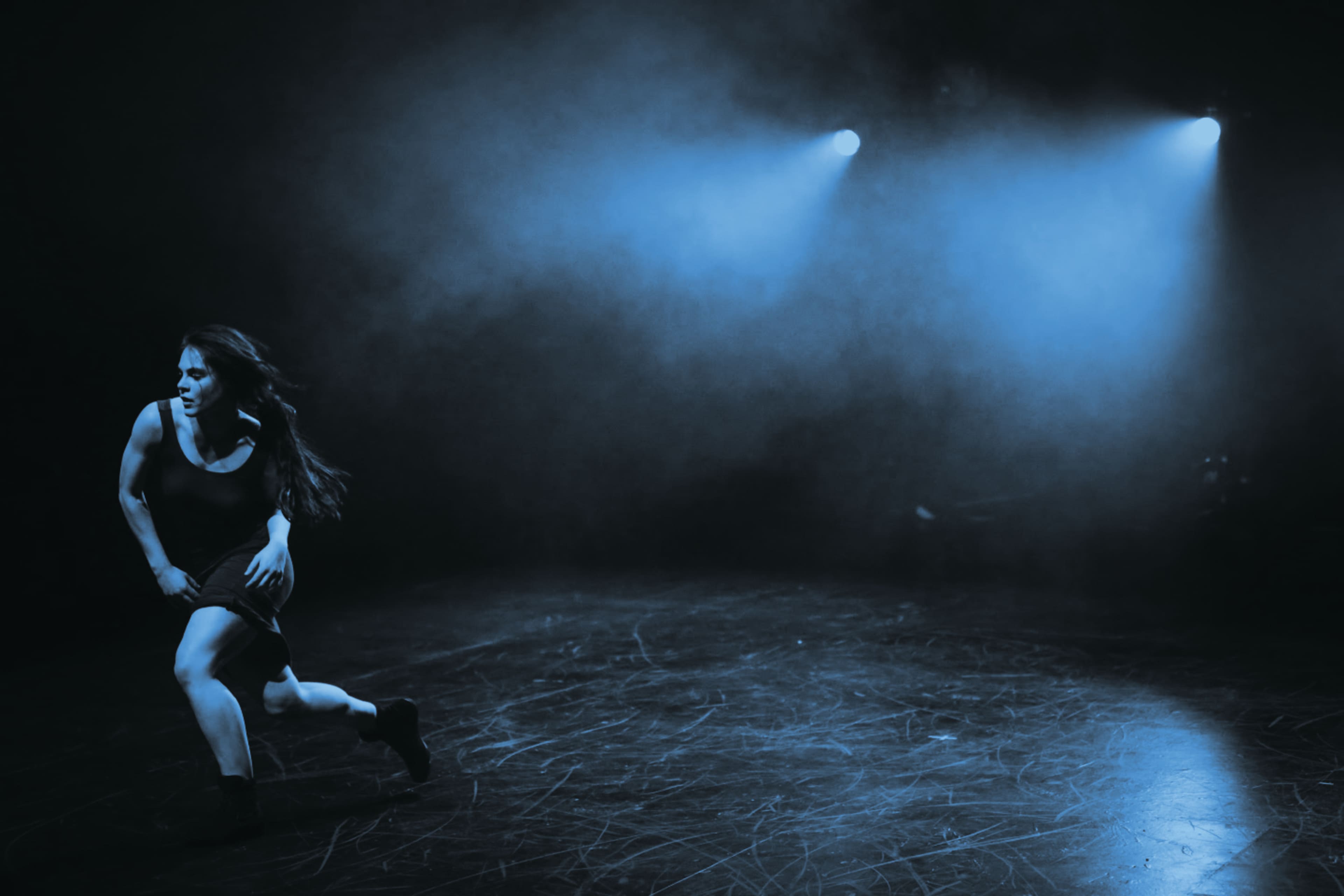 A dancer moves across a dimly lit stage, illuminated by blue spotlight beams amidst a smoky backdrop.