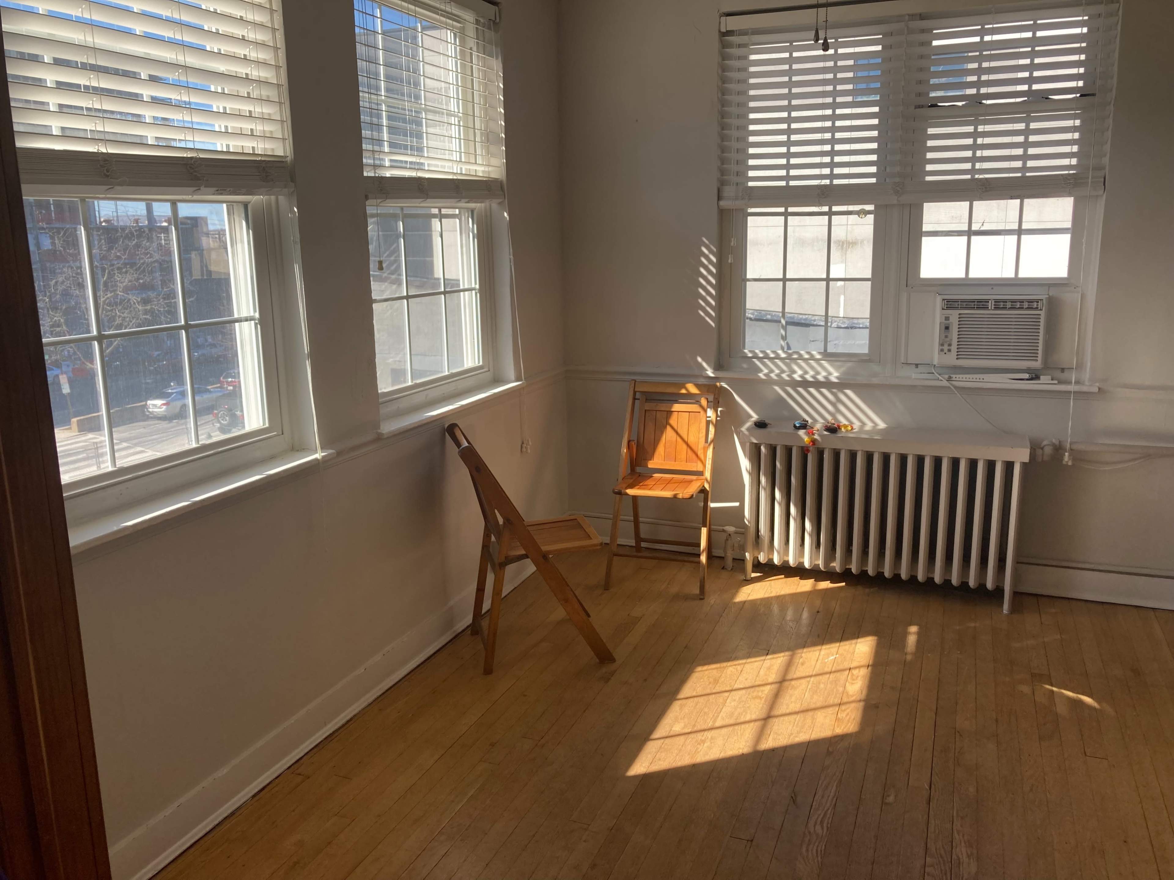 A bright, empty room with two windows, a folding chair, and an air conditioning unit below a radiator.
