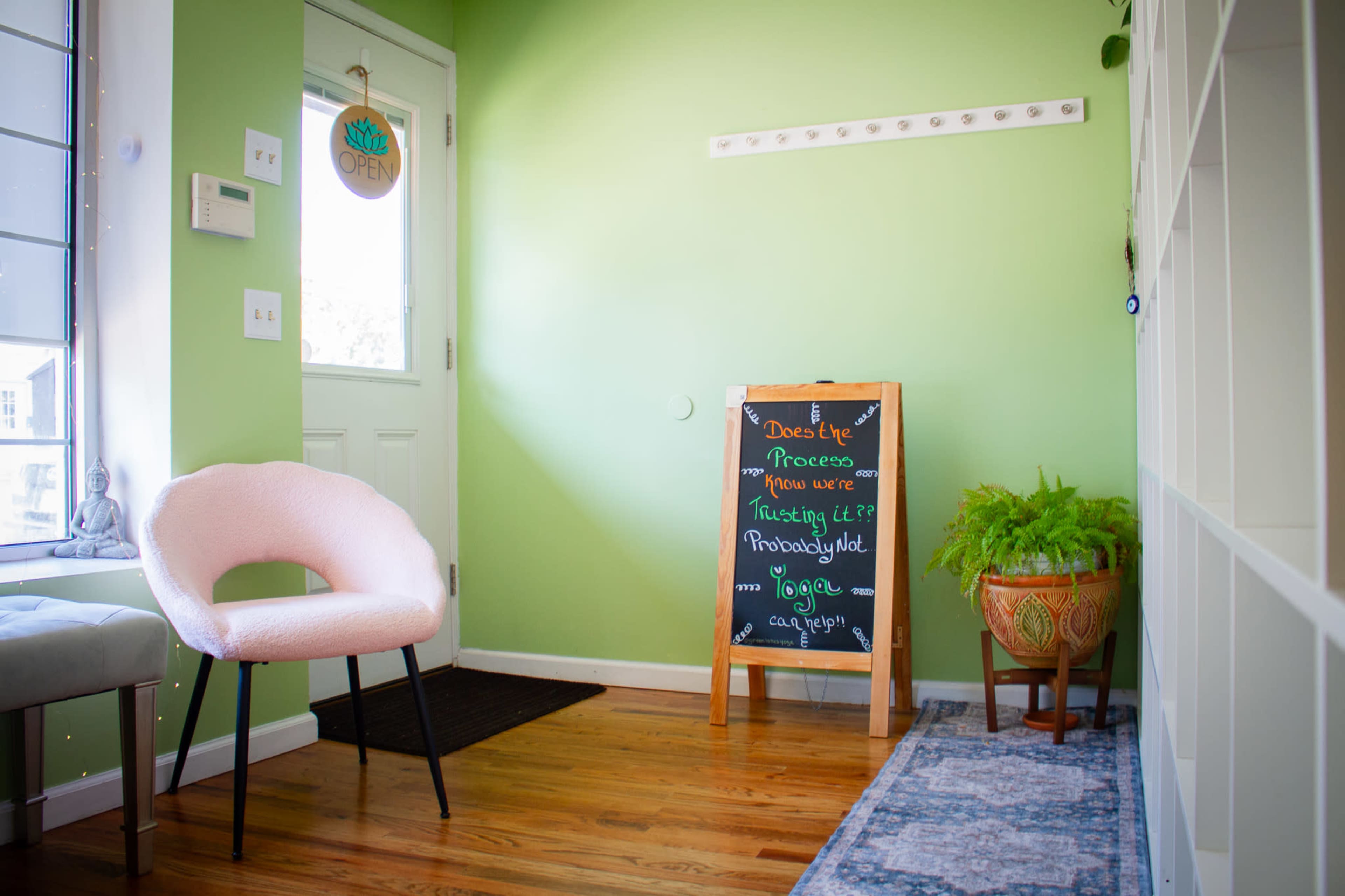 The image shows a light green entryway featuring a pink chair, a welcome sign, and a chalkboard with written information.