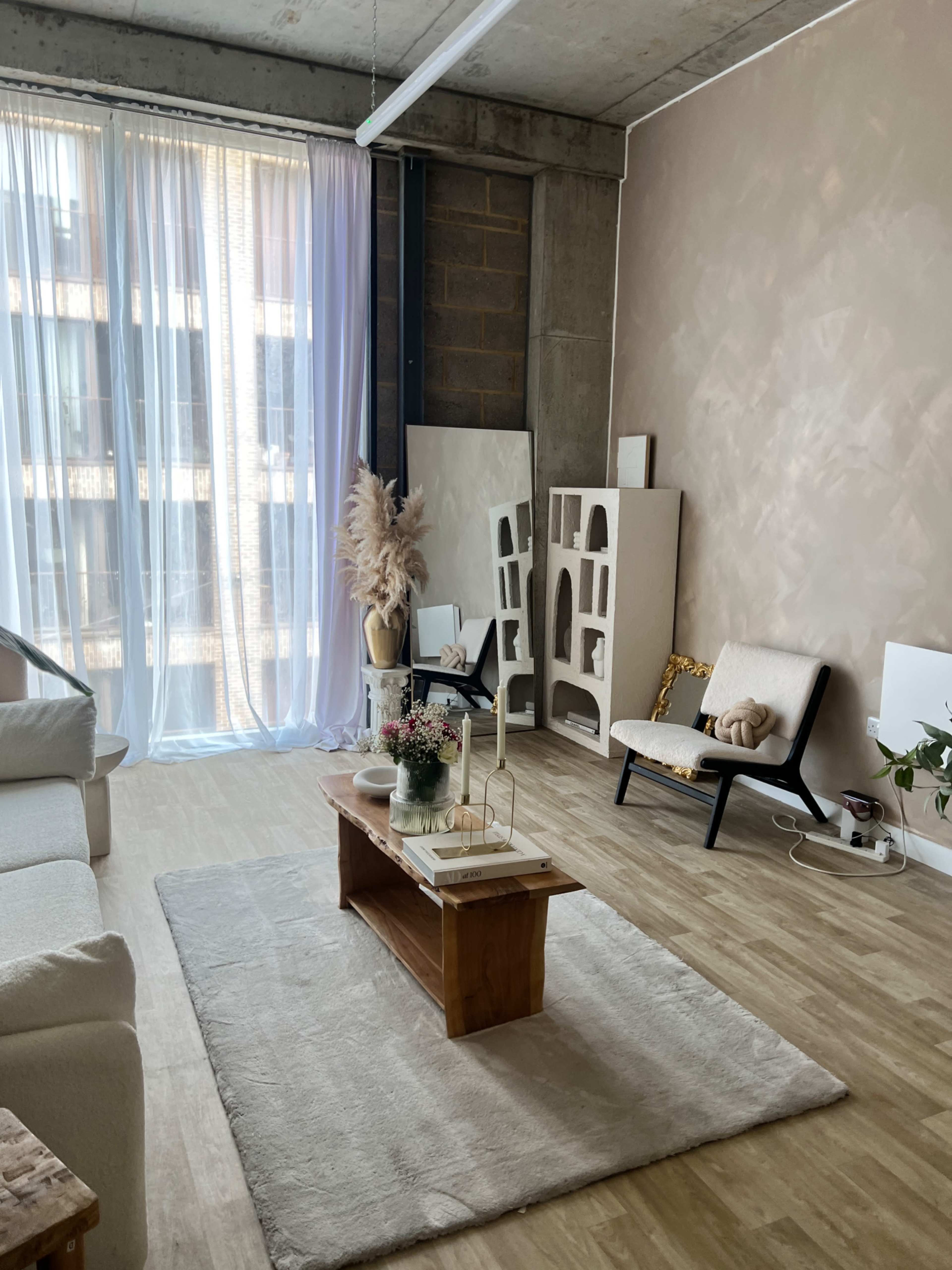 A cozy living room with a light-colored rug, a wooden coffee table, minimalistic furniture, and large windows covered with sheer curtains.