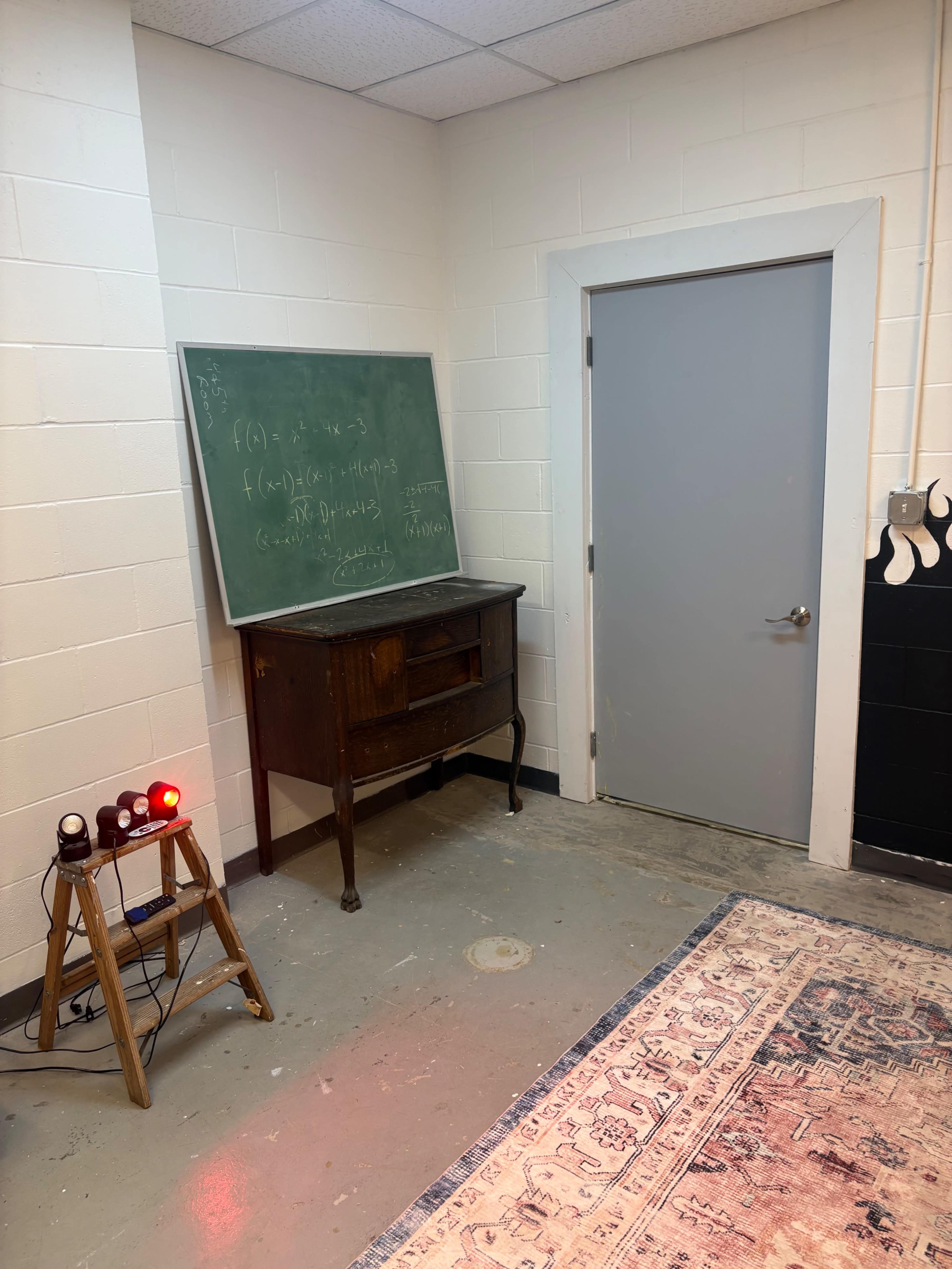 The image shows a corner of a room featuring a chalkboard with mathematical equations, a wooden cabinet, a closed door, and a rug on the floor.