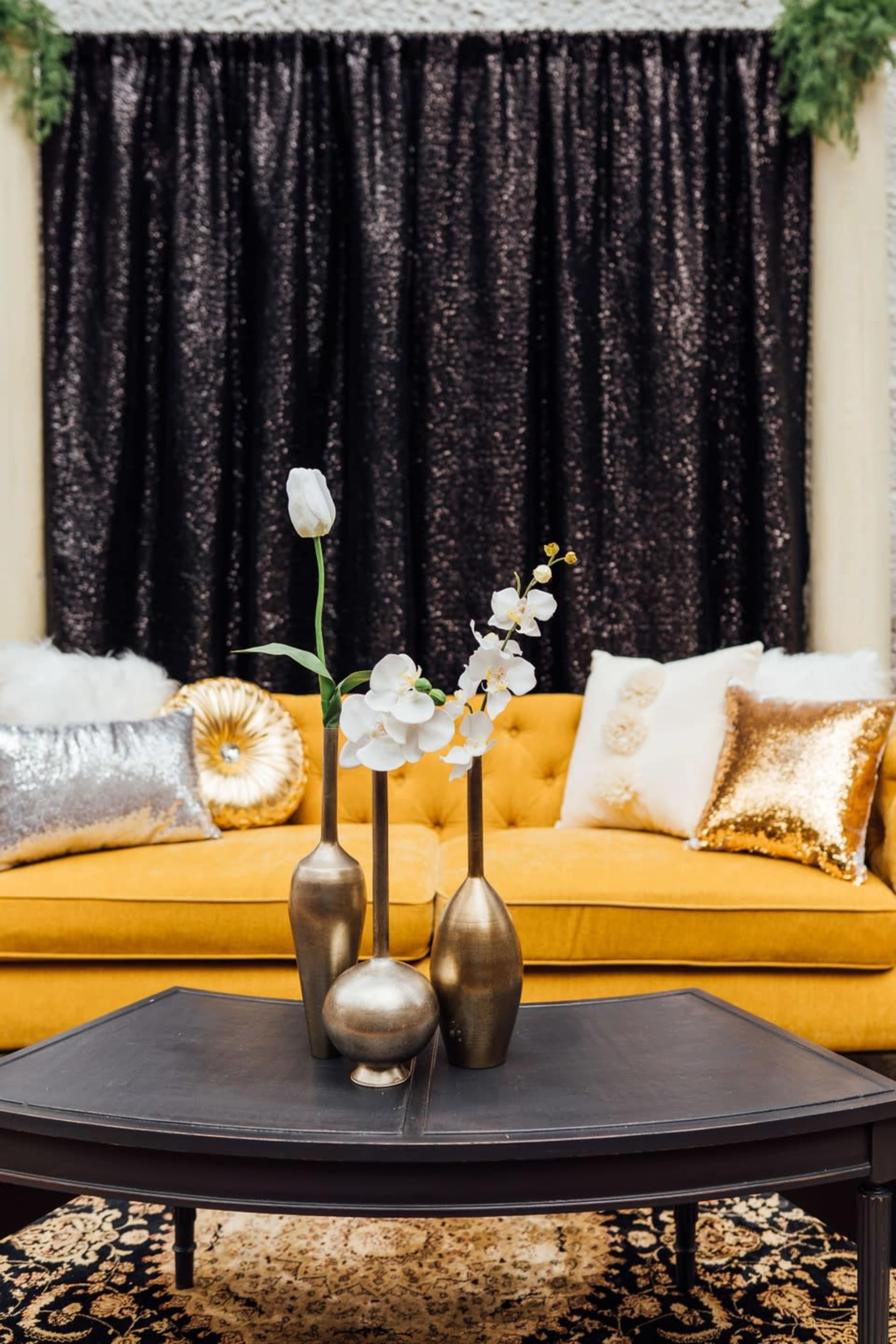 A yellow sofa is positioned in front of a black sequined curtain, adorned with decorative pillows and a coffee table featuring three gold vases with flowers.