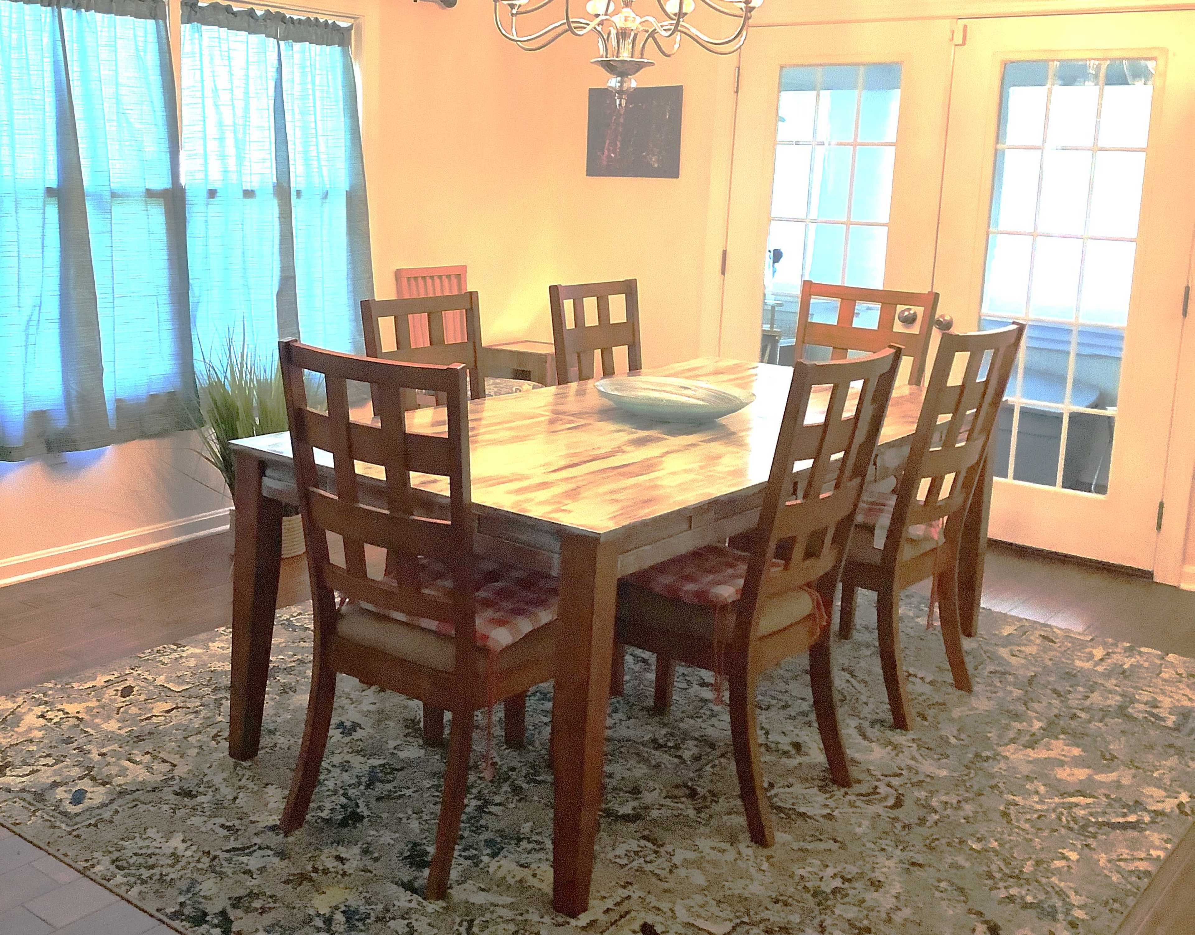 A wooden dining table with eight chairs is set in a well-lit room featuring large glass doors and blue curtains.