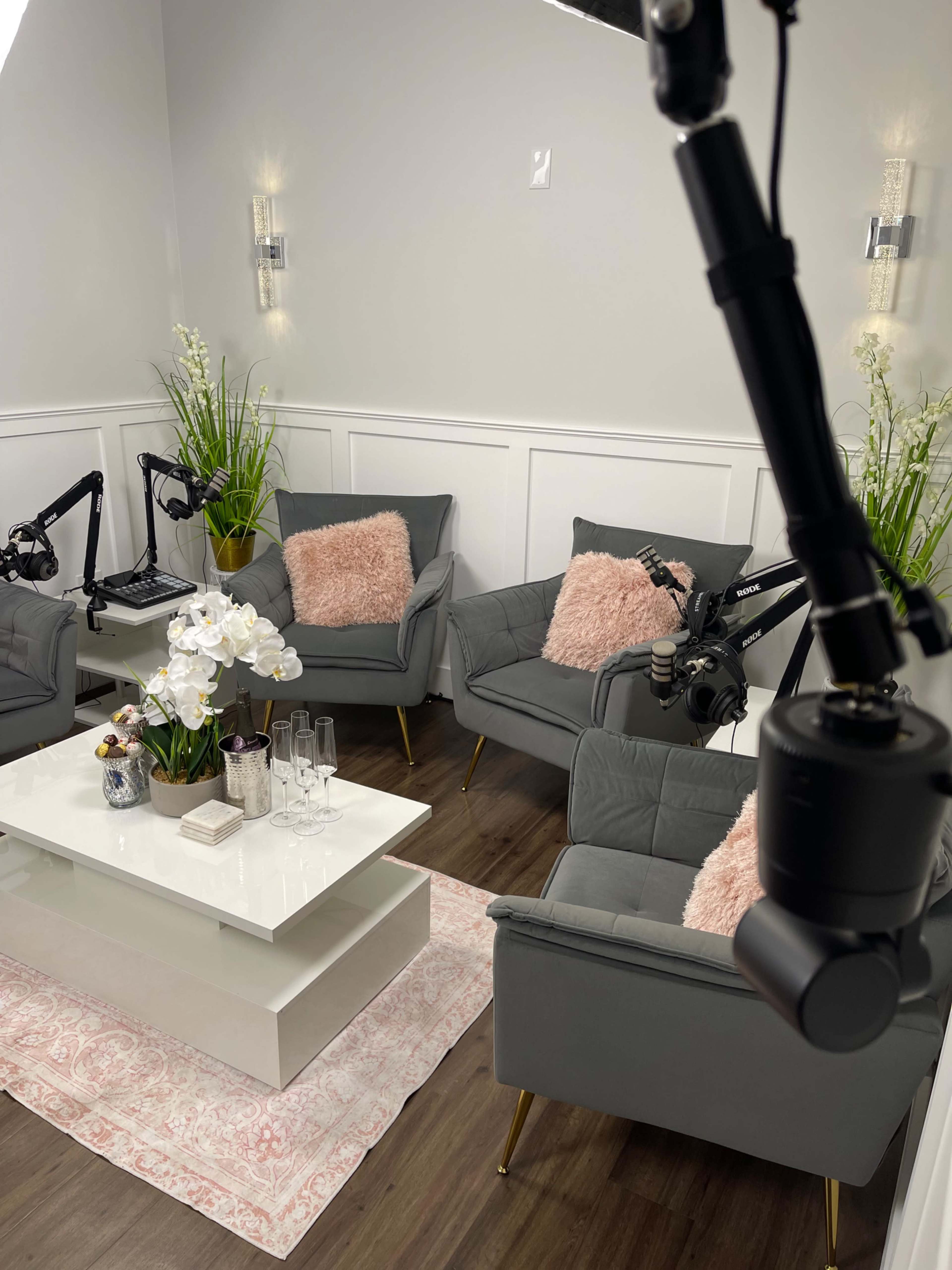 The image shows a stylish interview setup featuring two gray chairs with pink cushions, a white coffee table adorned with floral arrangements, and microphones positioned on stands.