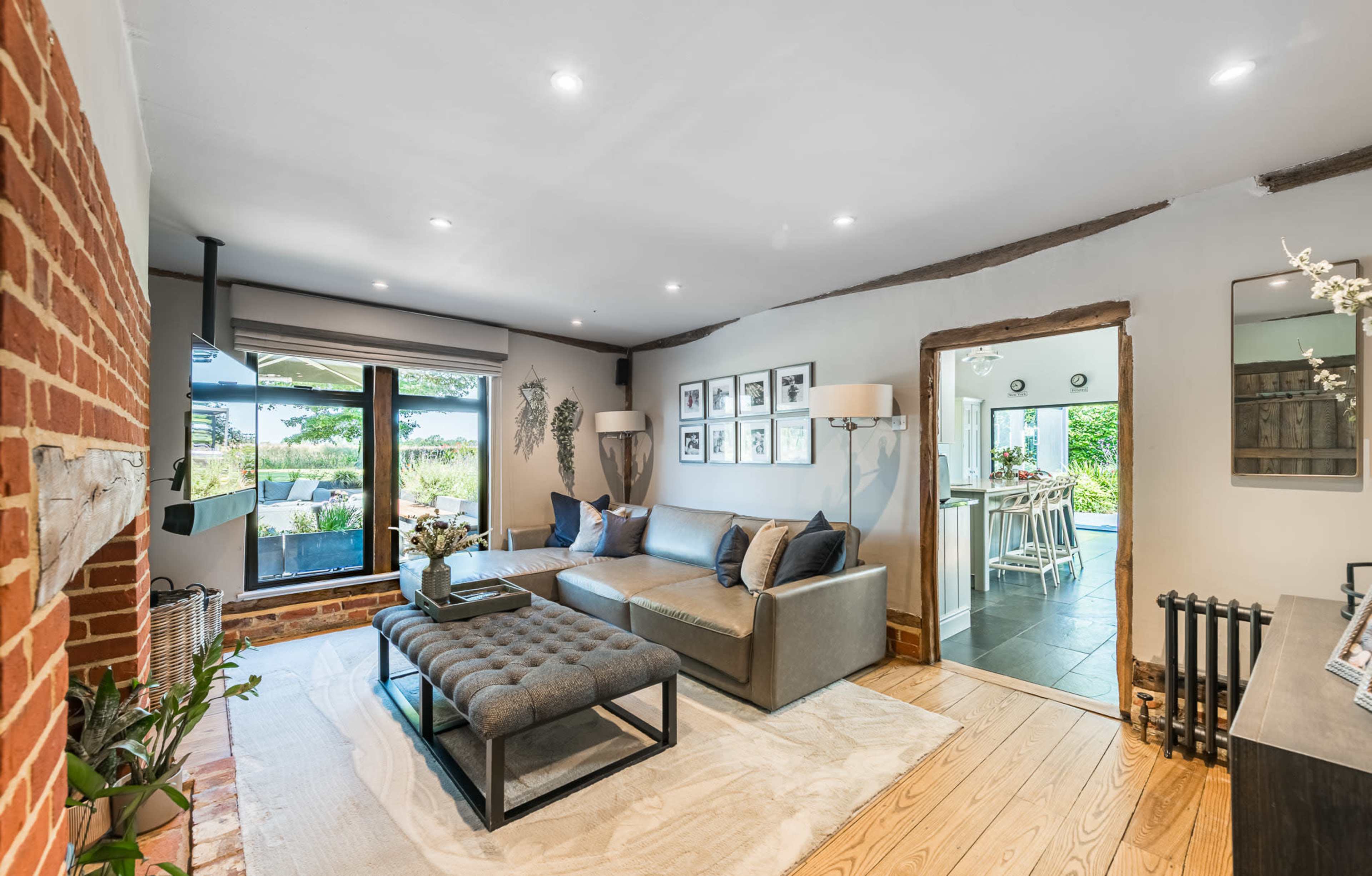 The image shows a modern living room with a gray sofa, a tufted coffee table, and large windows offering views of a garden.