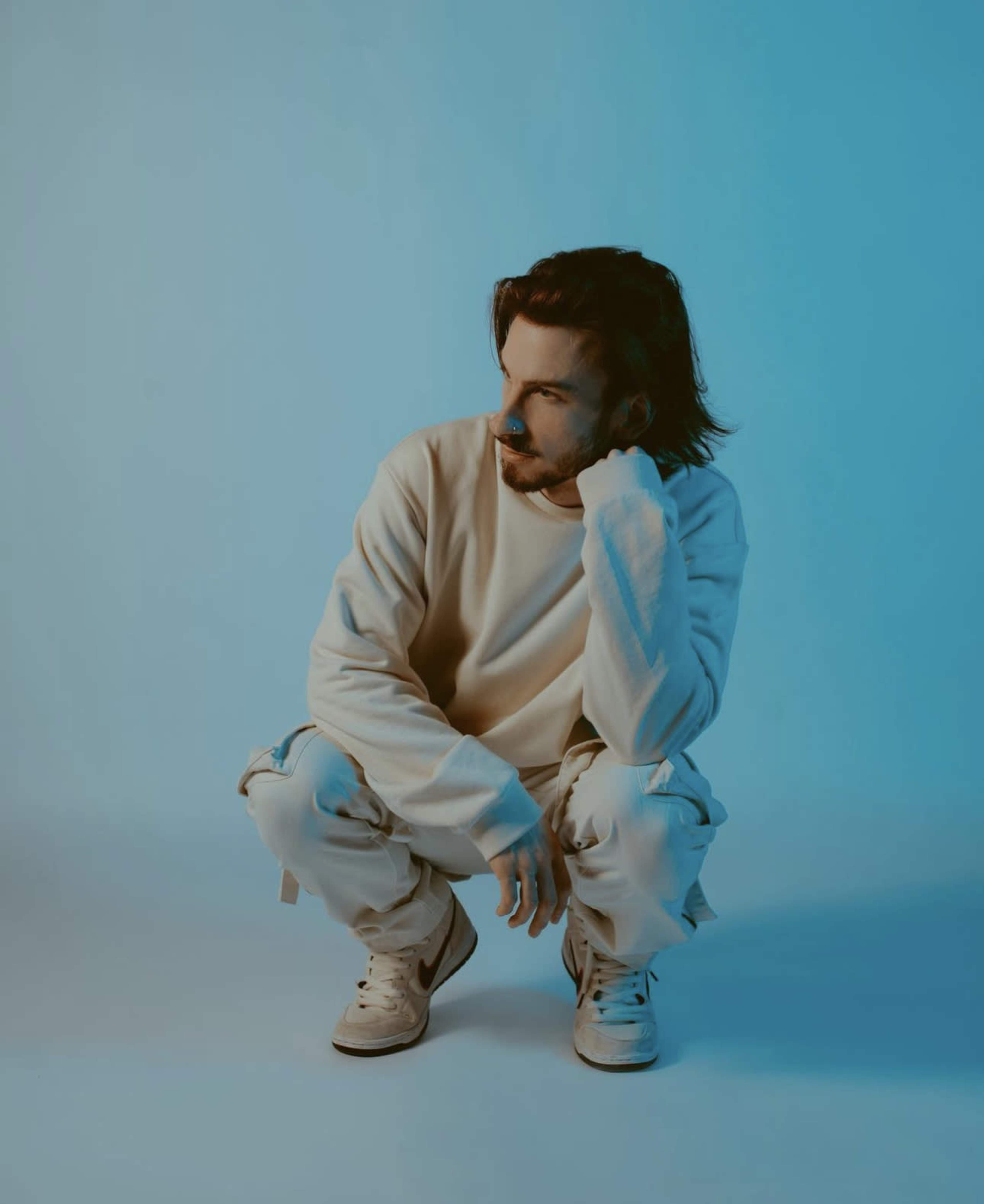 A person with long hair squats in a light-colored outfit against a blue backdrop.
