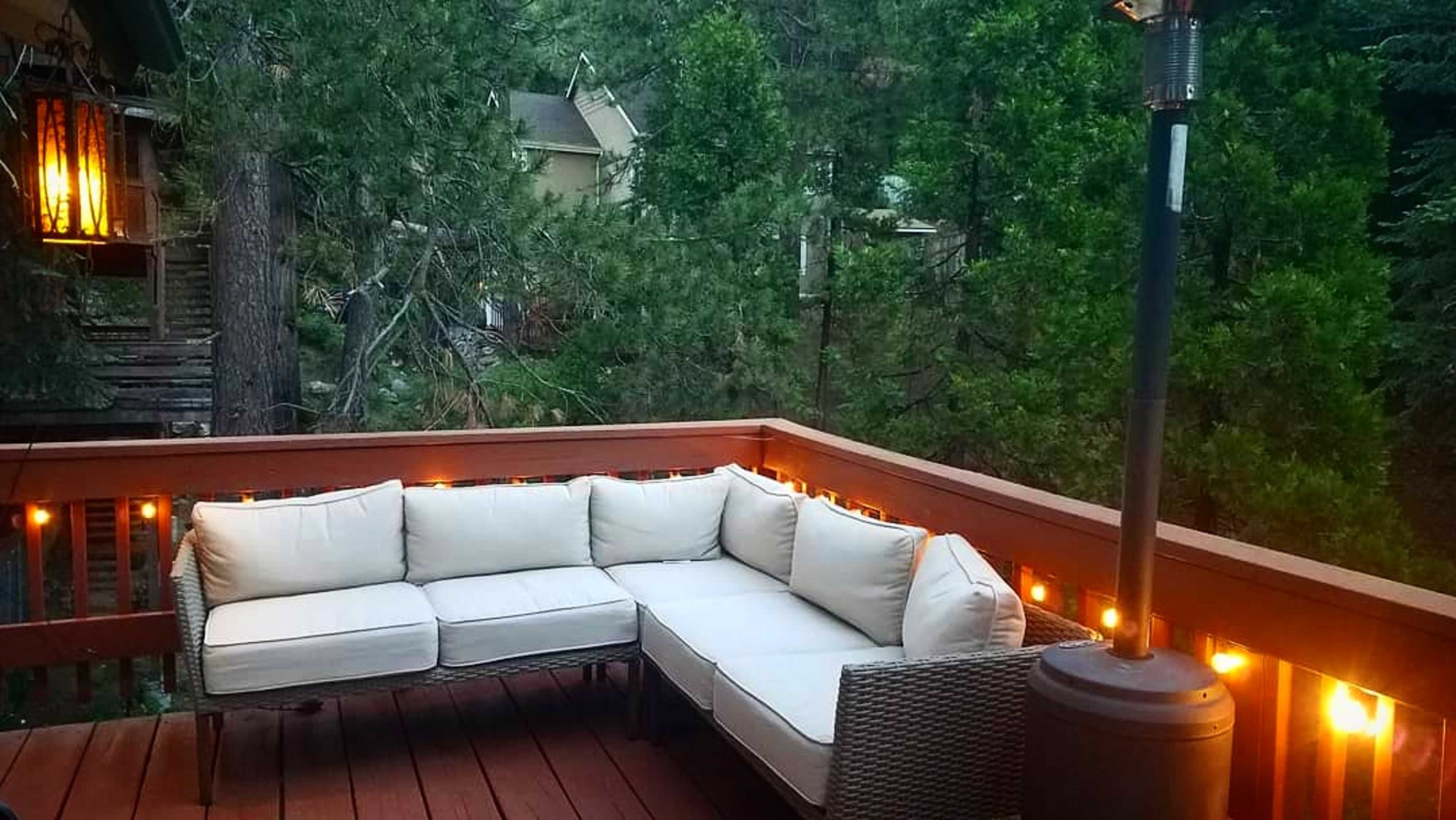 A corner sectional couch with white cushions sits on a wooden deck surrounded by tall trees and string lights.