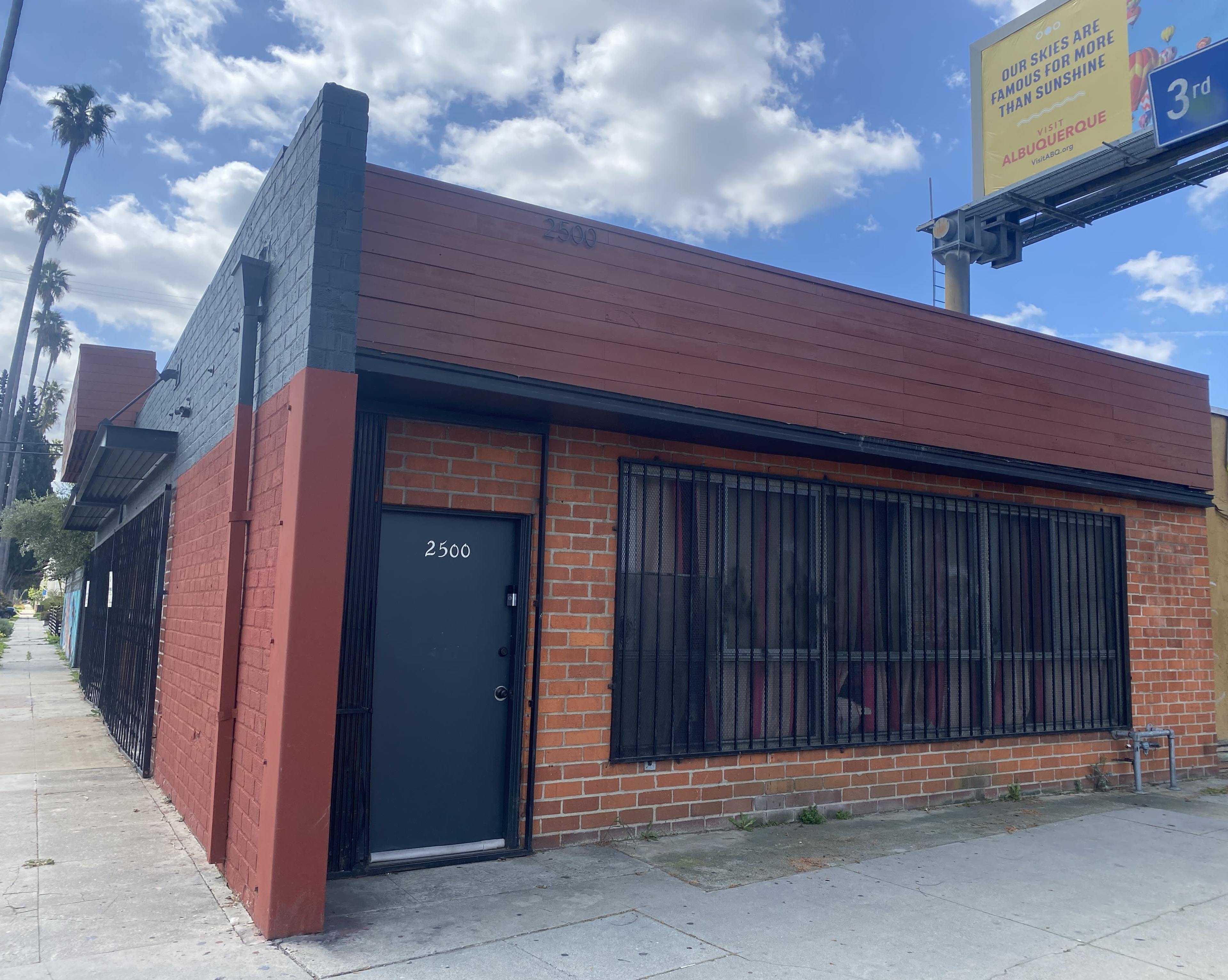 The image shows a single-story building with a red and black exterior and metal bars on the windows, located at the intersection of a street.