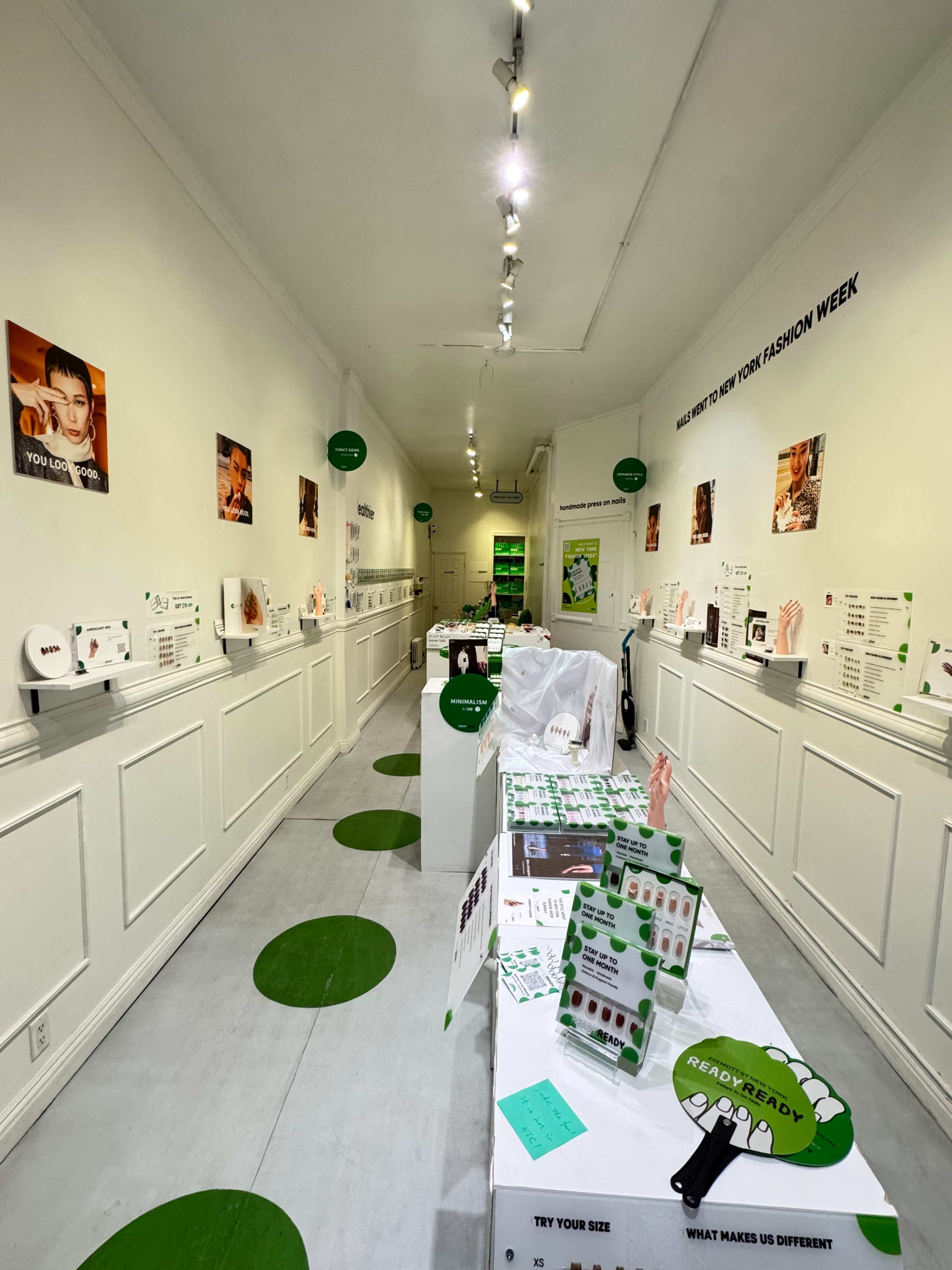 The image shows a narrow, white-walled exhibition space with green circular decals on the floor and various displays featuring fashion-related products and information along the walls.