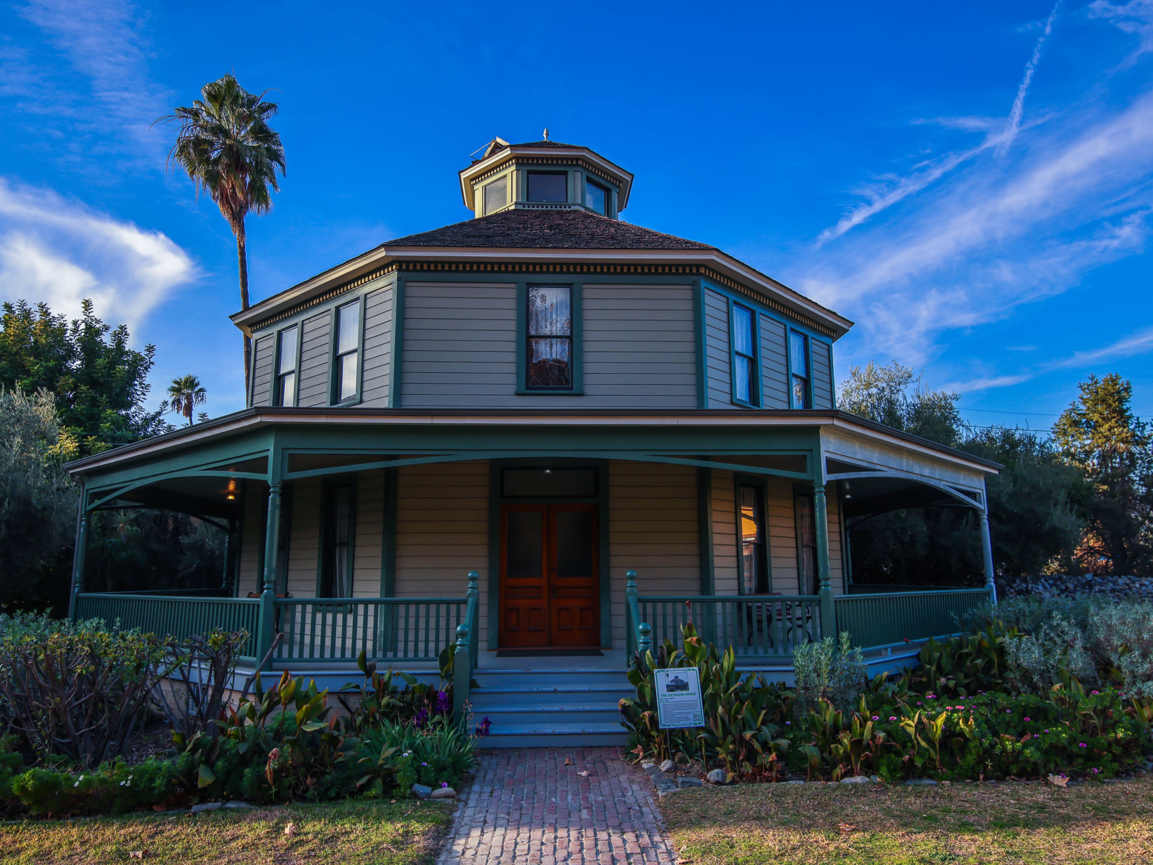 A large, round, two-story house with a green porch, surrounded by plants and trees under a blue sky.