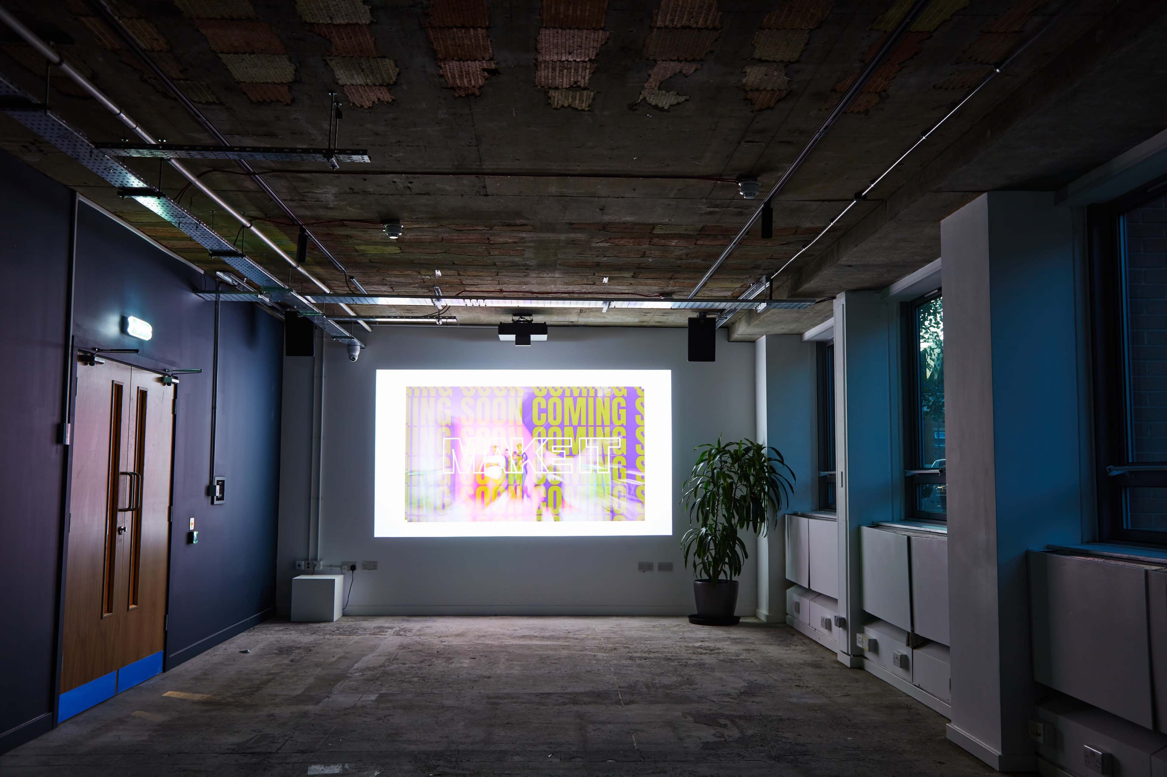 A brightly lit room features a large digital display on the wall with colorful text, and a potted plant sits in one corner.