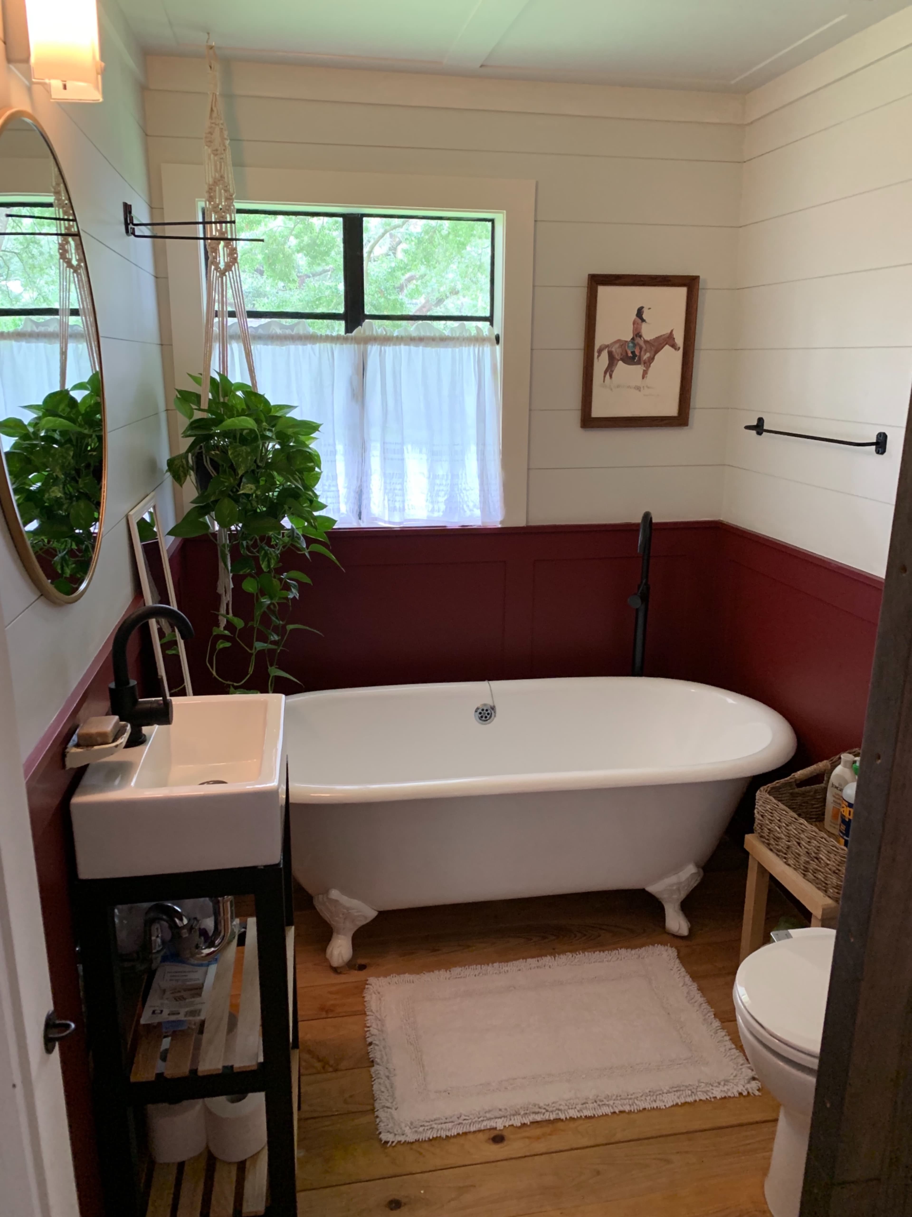 The bathroom features a freestanding white bathtub, a small sink on a black frame, and a large window with sheer curtains illuminating the space.