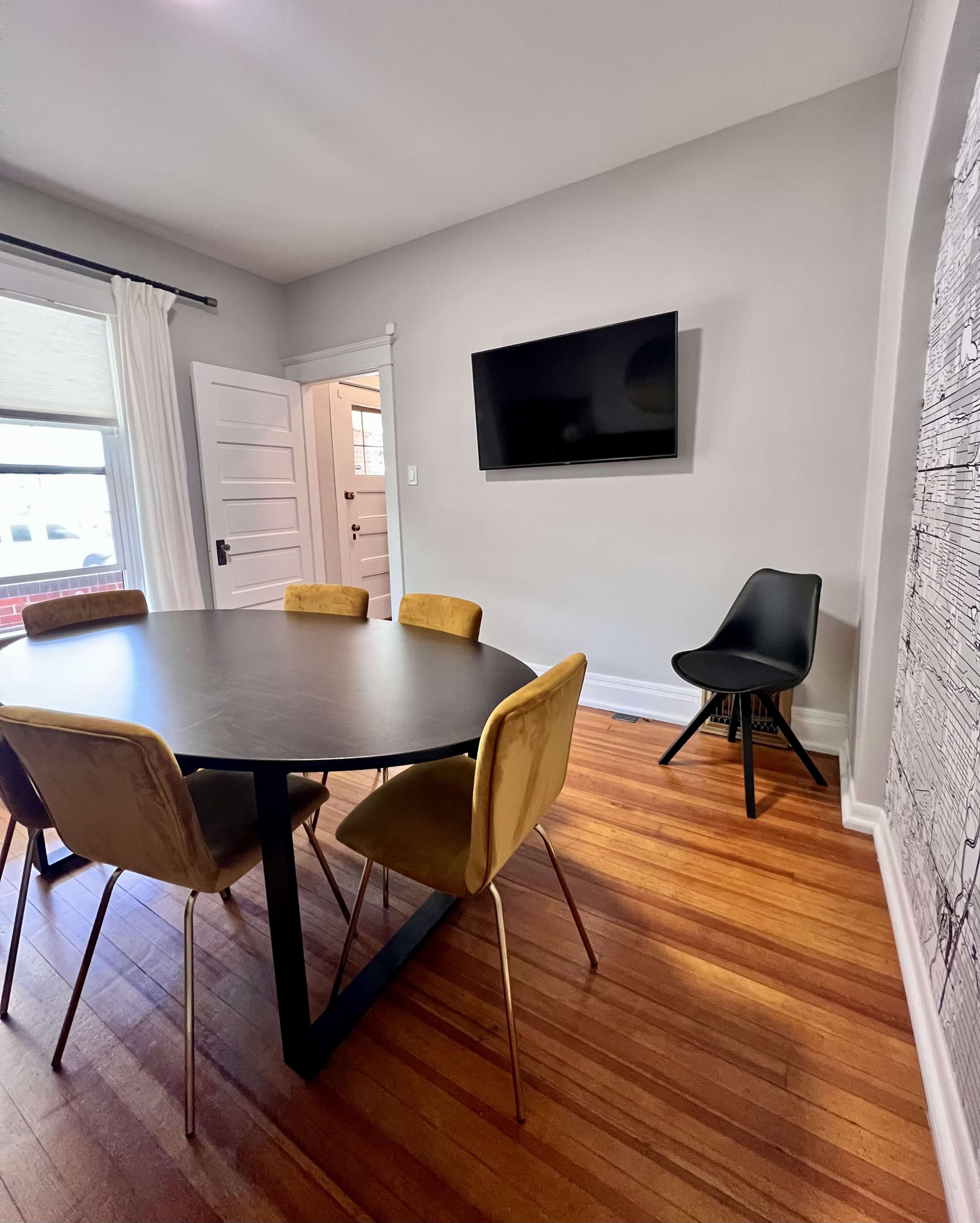 The image shows a dining area featuring a round black table surrounded by four golden upholstered chairs, a wall-mounted television, and a doorway to another room.