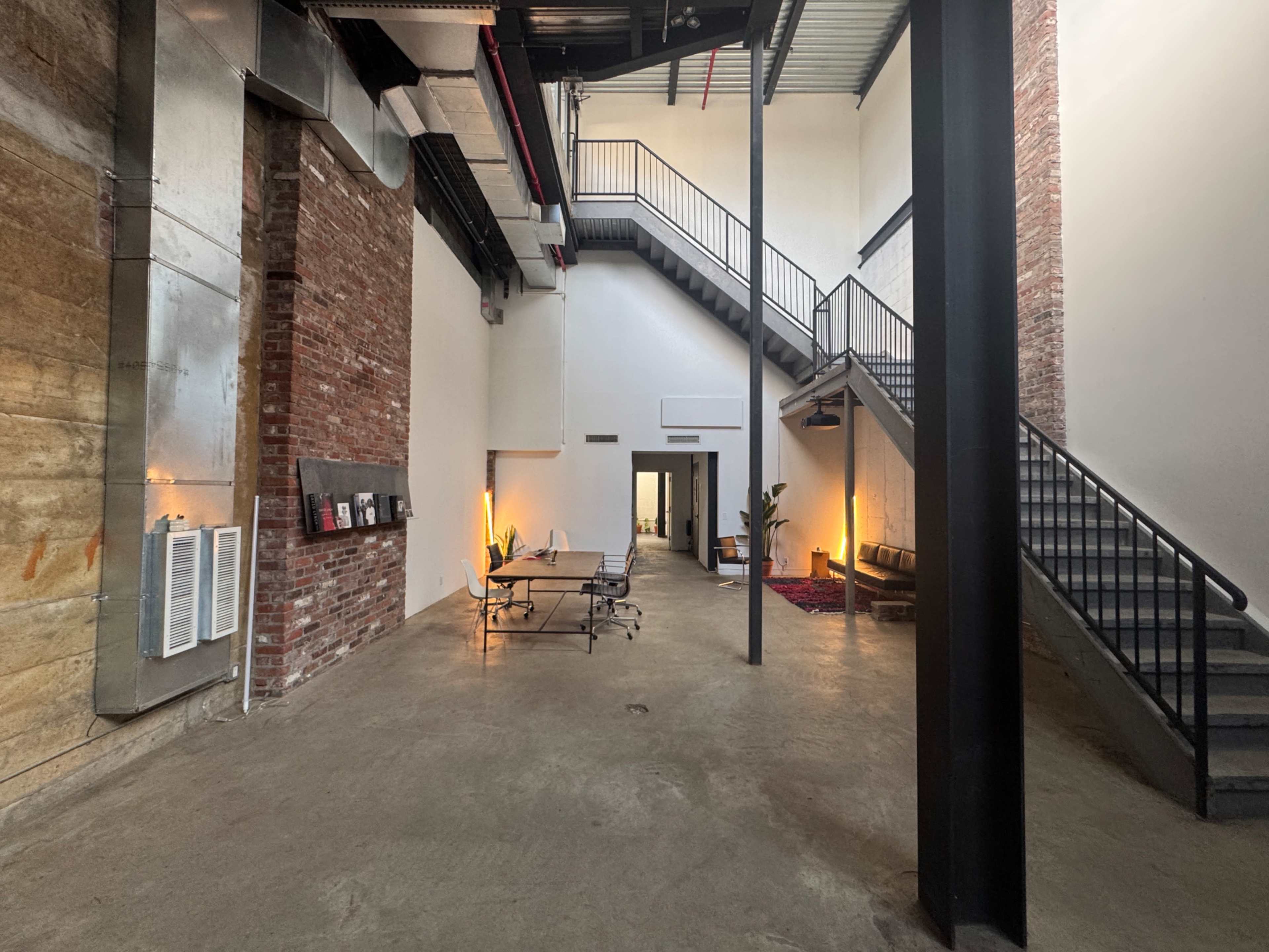 The image depicts a spacious, industrial-style interior with exposed brick walls, concrete flooring, and a staircase leading to an upper level.
