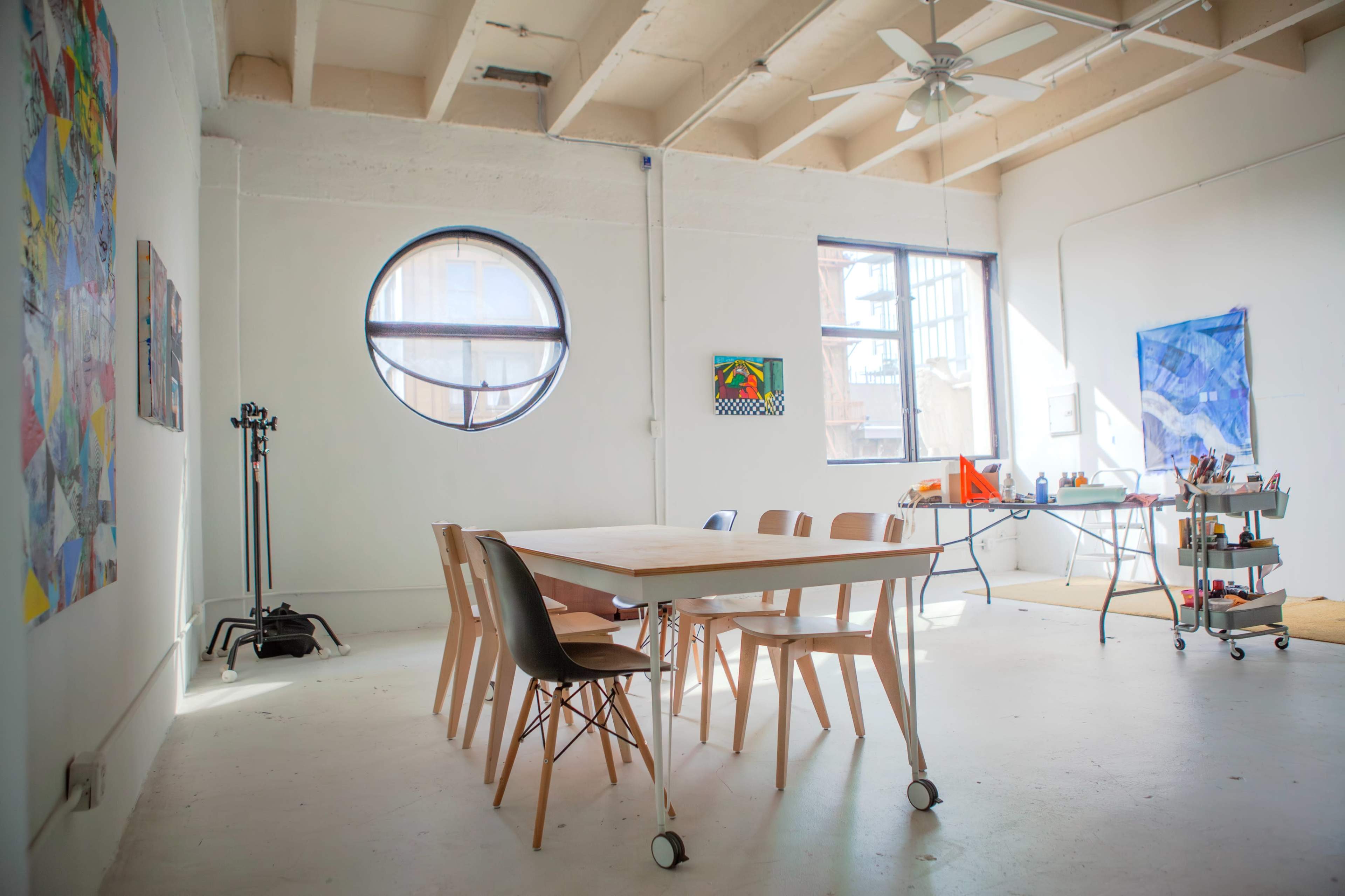 A spacious, well-lit room features a large table surrounded by wooden chairs, with artwork on the walls and a worktable in the corner.